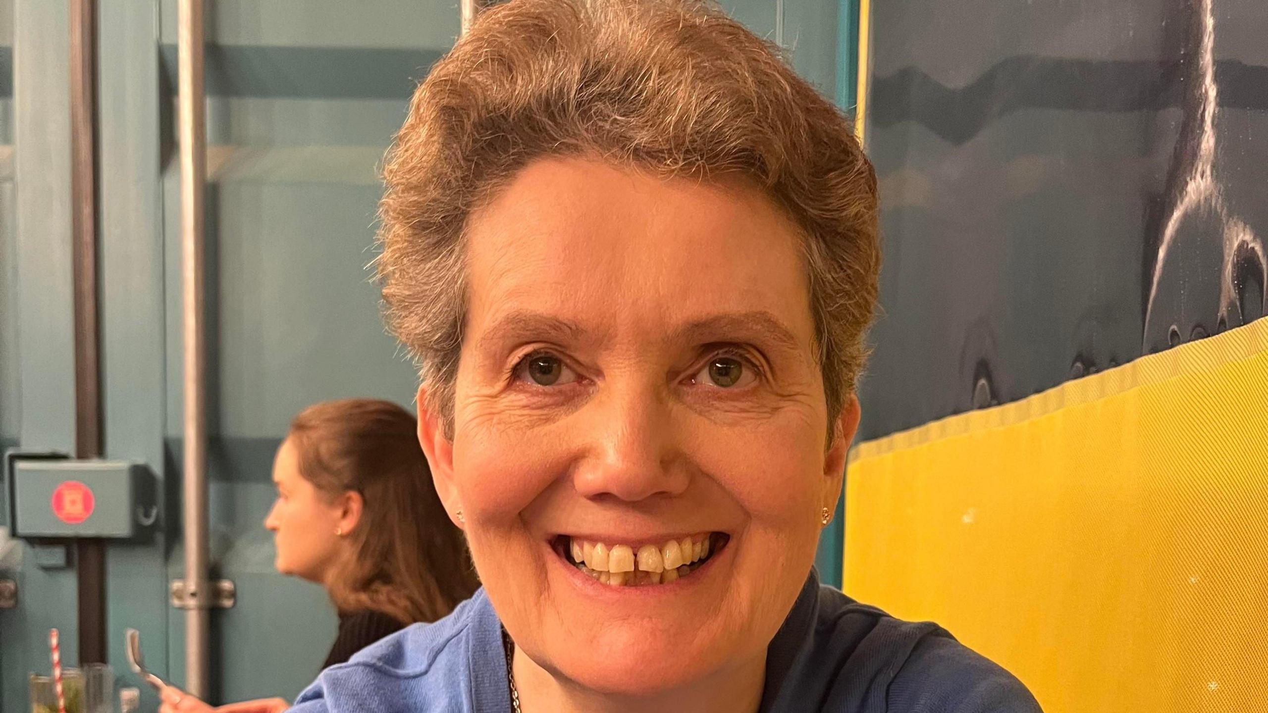 Kim is sat in a cafe, smiling at the camera. She has short, wavy grey hair. She is wearing a blue knitted top and necklace. The shot is of her head, and you can see someone sat behind her.