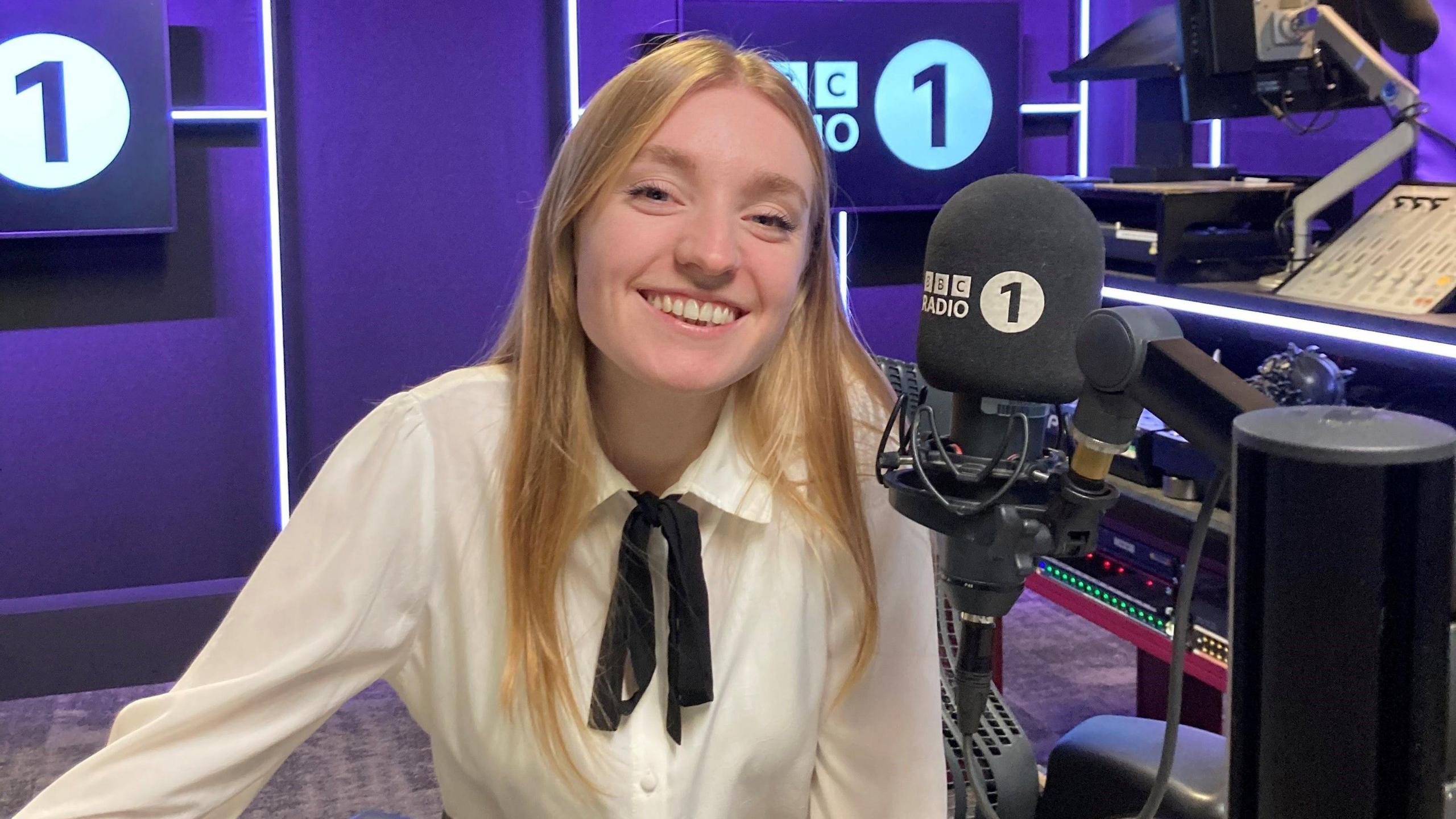 Kitty Perrin, who has long, blonde hair and is wearing a white shirt, looks directly at the cameral as she sits in front of a Radio 1-branded microphone. The shot is in a radio studio with purple walls. The BBC Radio 1 logo is on the walls. 