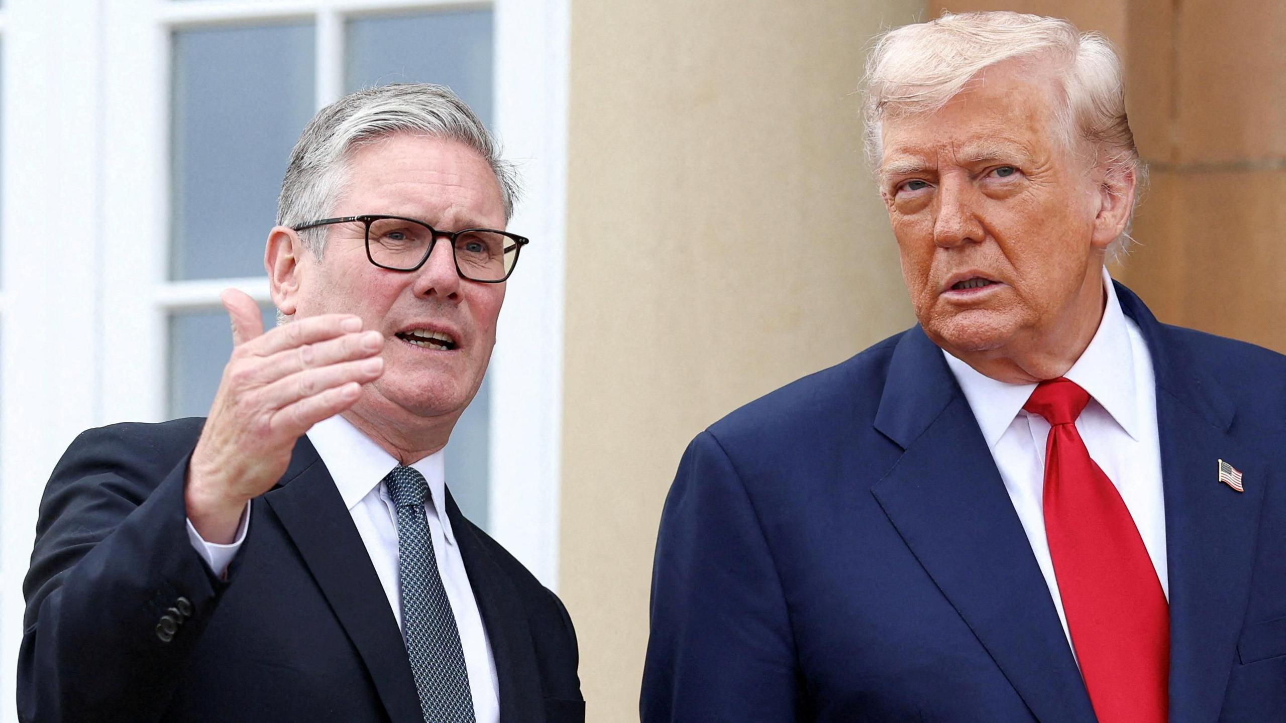 Sir Keir Starmer and Donald Trump outside a sandstone building. Starmer has raised his right hand and Trump is listening to what he is saying. Both men are wearing suits, ties and white shirts.