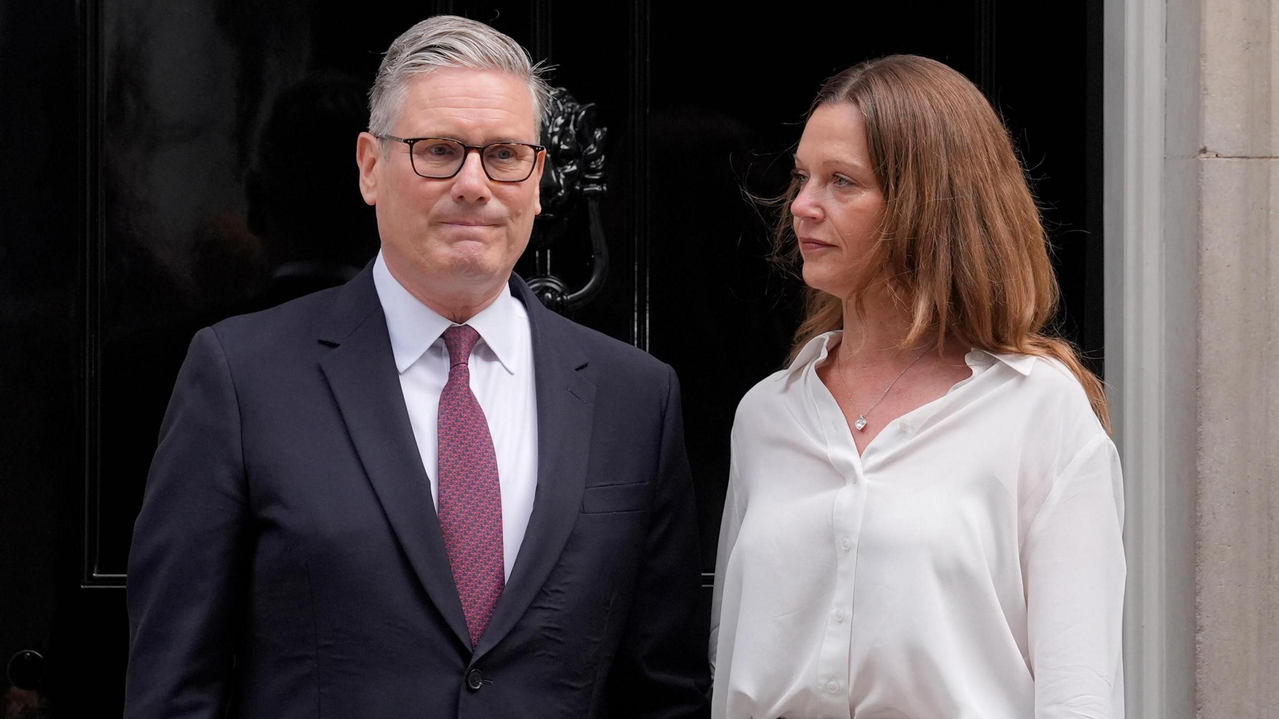 Sir Keir Starmer stood with his wife Victoria Starmer outside the door of No.10 Downing Street. The door is dark black. Starmer is wearing a suit, a white shirt, and a red tie. His wife is wearing a white shirt.