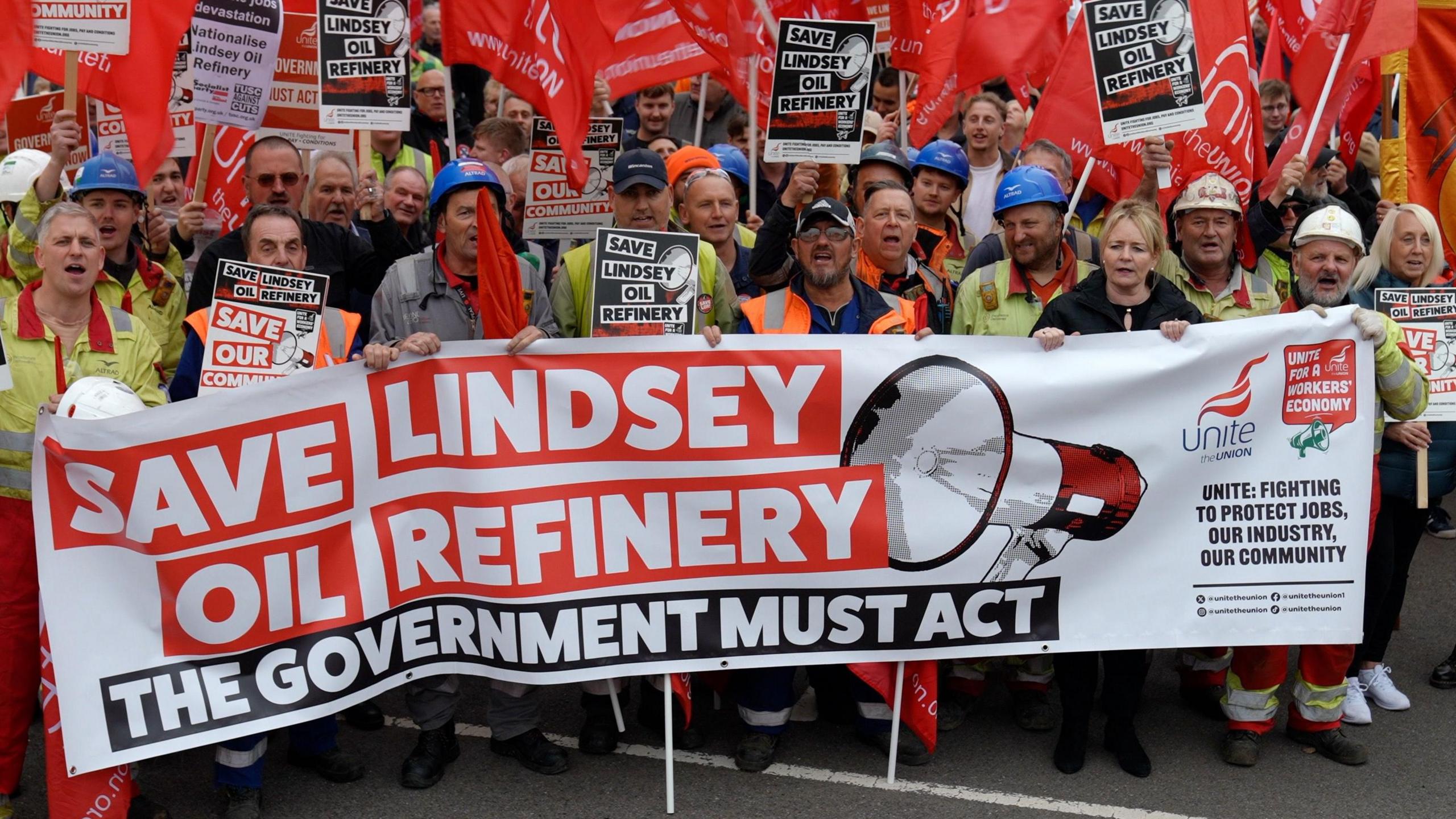 Dozens of workers and union reps, the majority men and most wearing hi-vis jackets and helmets, chant and wave red flags and signs as they stand behind a large white banner. On the banner, words read "SAVE LINDSEY OIL REFINERY" in white letters on a red background. Another sentence reads "THE GOVERNMENT MUST ACT" in white letters on a black background. The banner also includes a drawing of a megaphone and the logo of the Unite union.