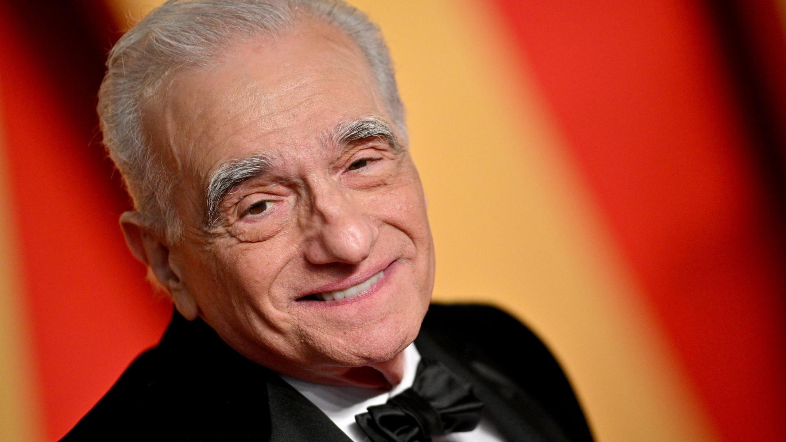 Martin Scorsese is an older man with white hair. He is smiling as he poses for a picture at a film event against a background of red and orange colours. He is wearing a tuxedo. 