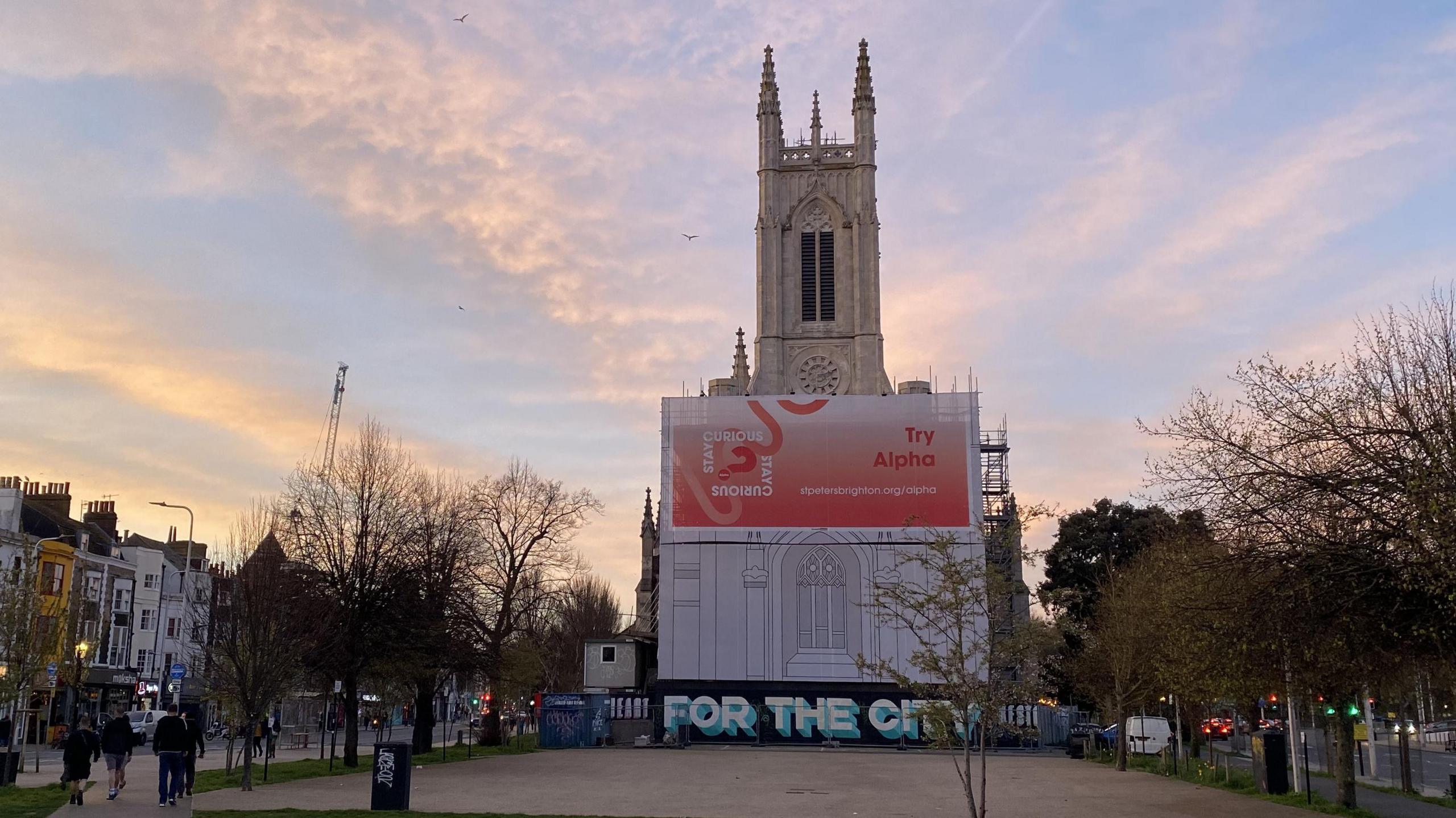 A church. The lower half of the church is wrapped in scaffolding and sheets. The sun is going down in the background.