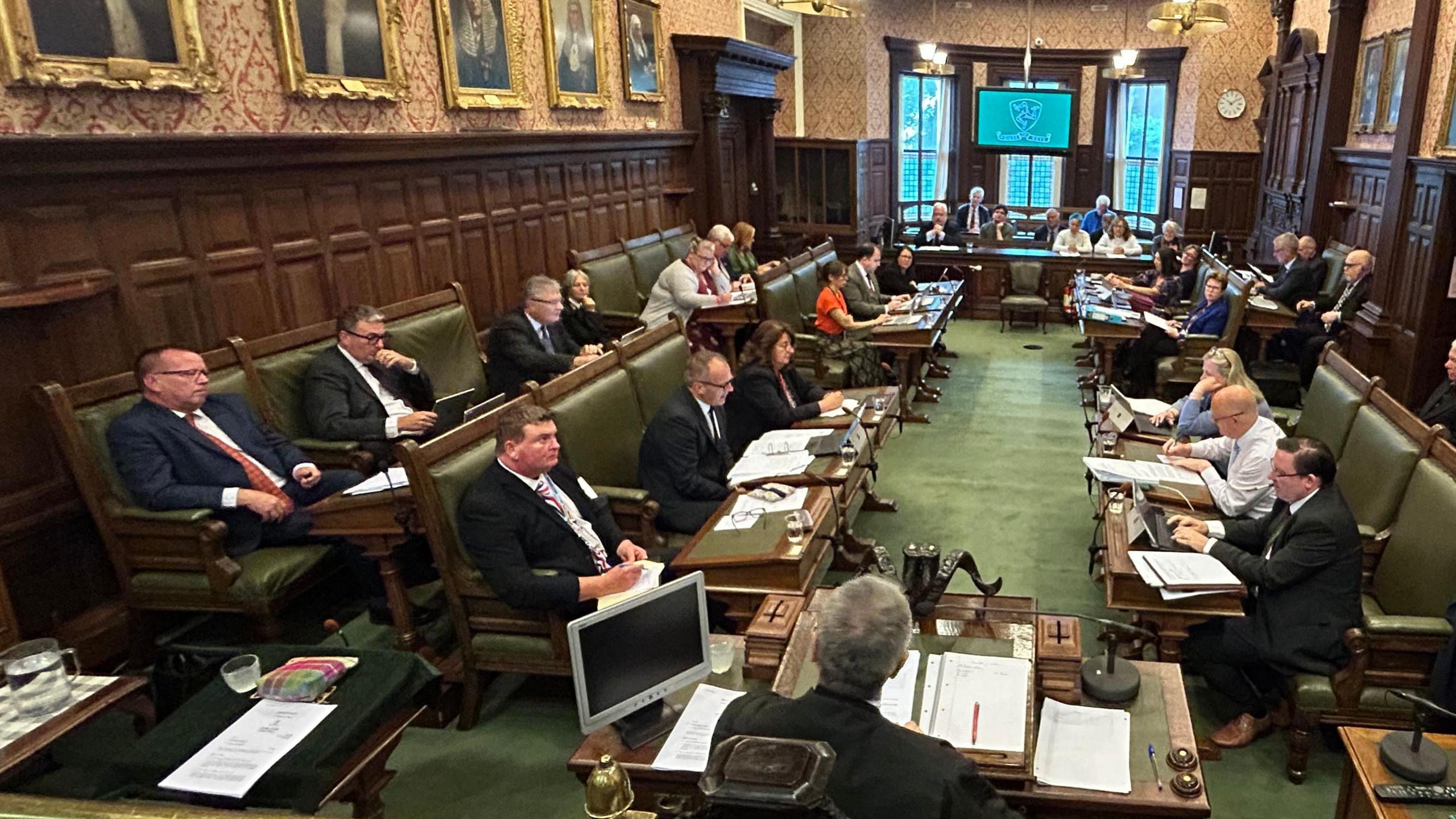 The inside of the House of Keys chamber, which has green carpet, wooden panels on the walls and large pictures in gold frames. Politicians, male and female, sit alongside each other on green leather chairs, with desks in front of them. Members of the public watch on from a gallery. Other members of the House of Keys sit nearby and listen on. The seats and benches are wooden and upholstered with avocado green leather.