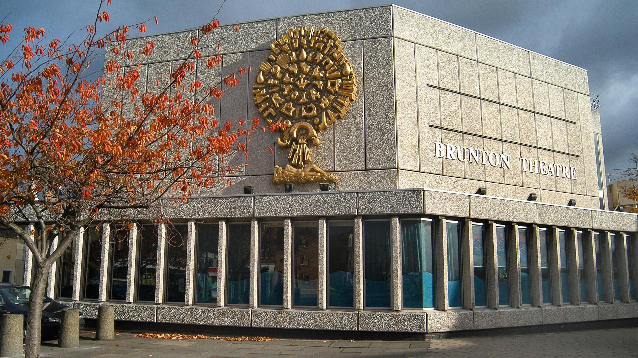 A side view of the exterior of Brunton Theatre. The building is grey, with a gold design on one side. The words Brunton Theatre are on the other side in white. A tree with red and orange leaves is in front of the building above three stone bollards.