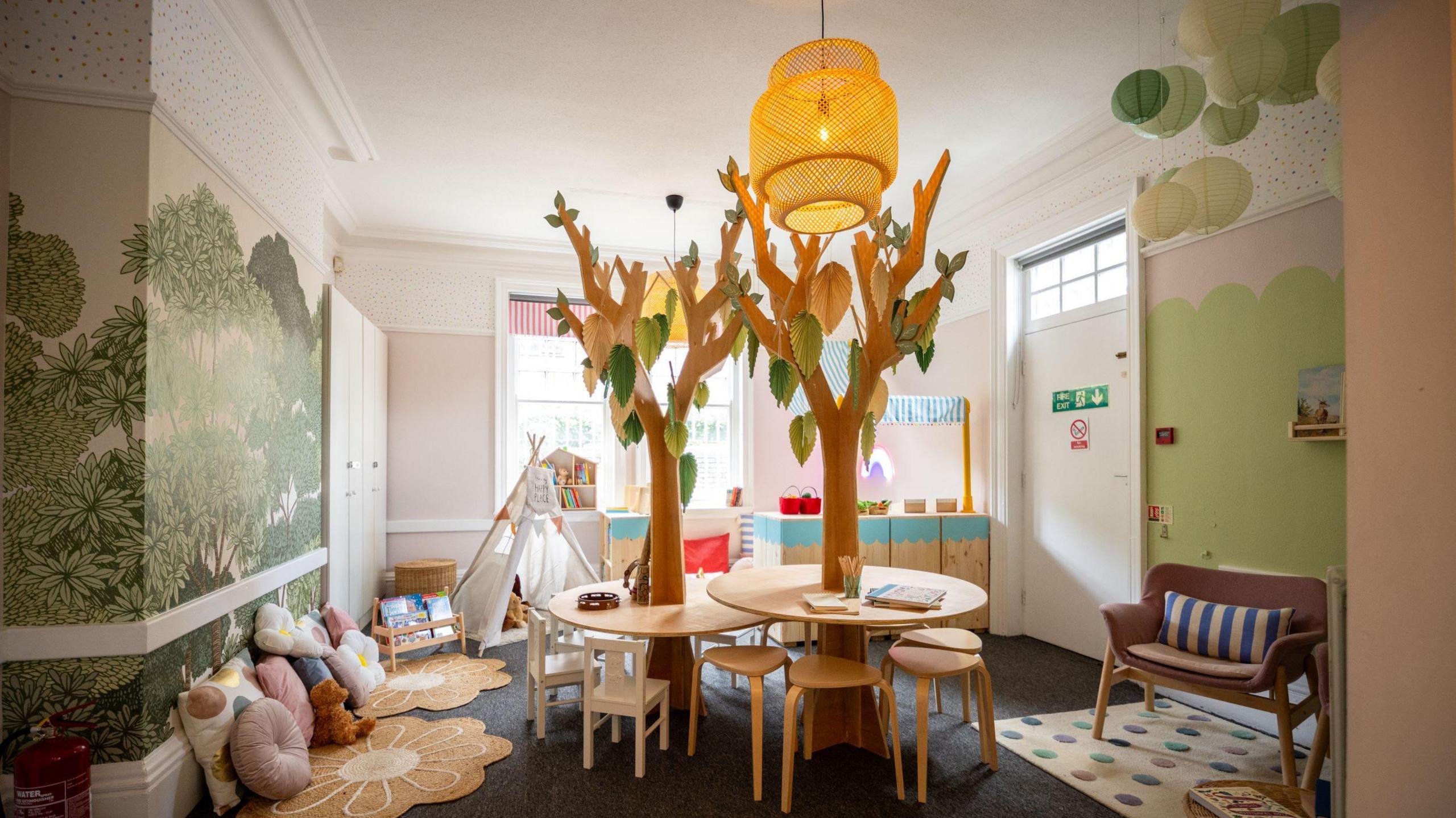 A room with green forest-themed walls. In the centre are wooden tables with fake trees risin up through the middle. To the left of the room there are cushions lining the floor