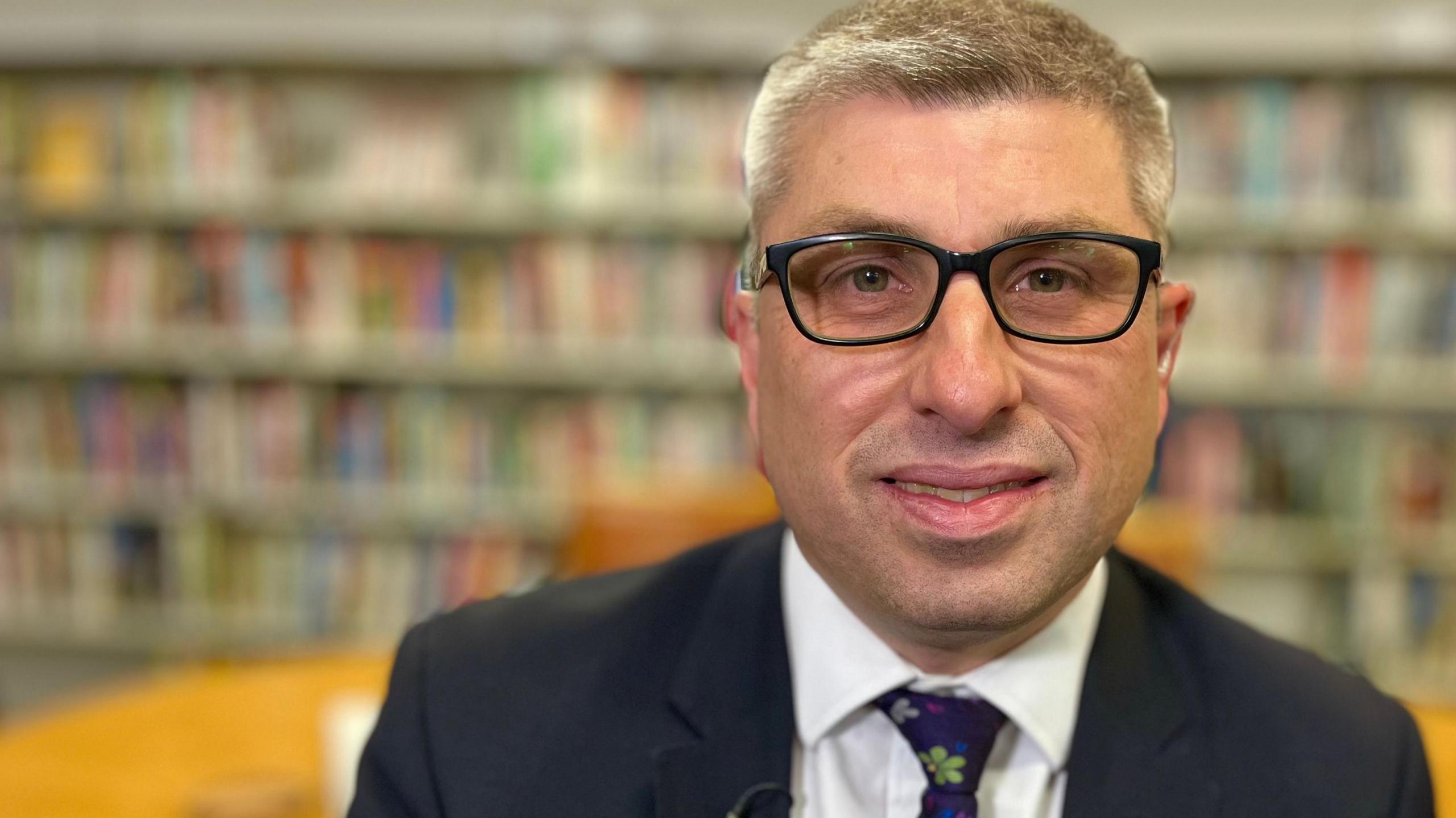 Mark Arnull is in the middle of the image looking towards the camera. He has short grey hair and is wearing black framed glasses. He is wearing a navy suit jacket, white shirt and a purple tie. In the background are book shelves.