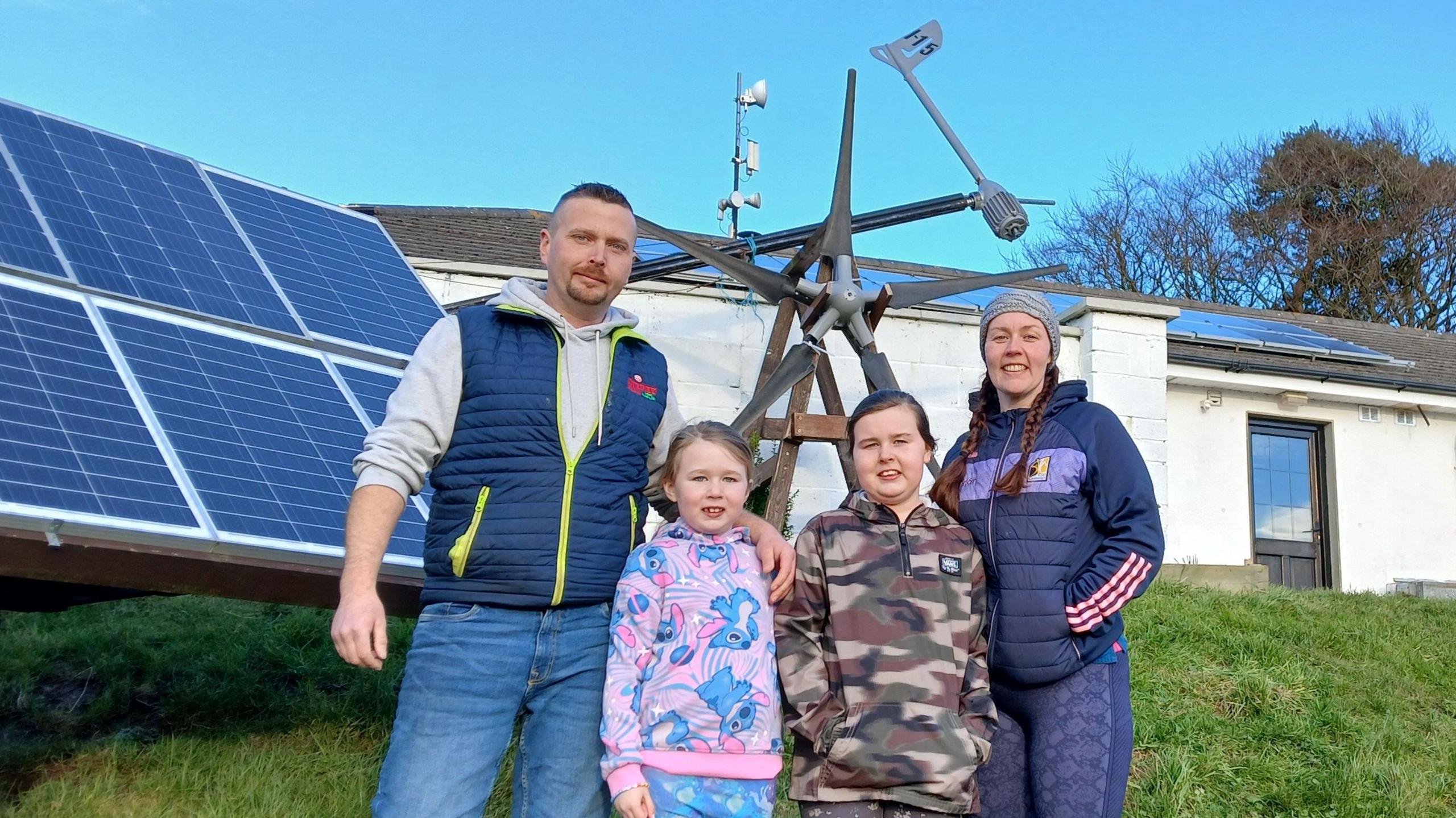 Michael Wilkinson with his wife and two daughters, standing outside their home in County Kilkenny.  Two dogs are in the background in their tiered garden.  There are solar panels on the roof and propped up on the lawn.  There is also a wind turbine behind them. 