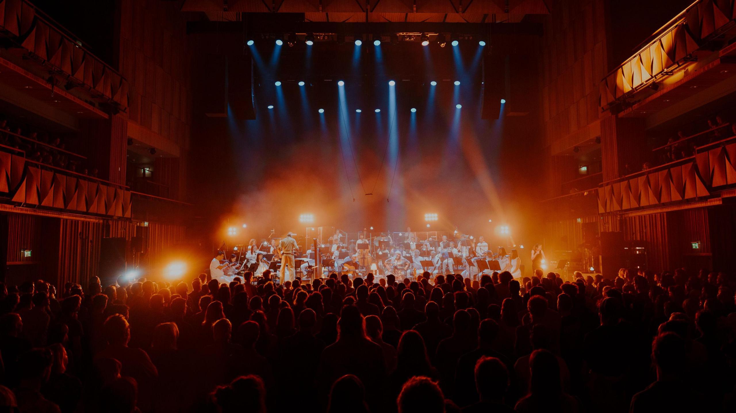A photo taken from the back of a crowd looking at the stage at the Bristol Beacon during the BBC Proms. The crowd are in darkness although you can see their heads, and on the stage the Paraorchestra are performing, illuminated by bright lights