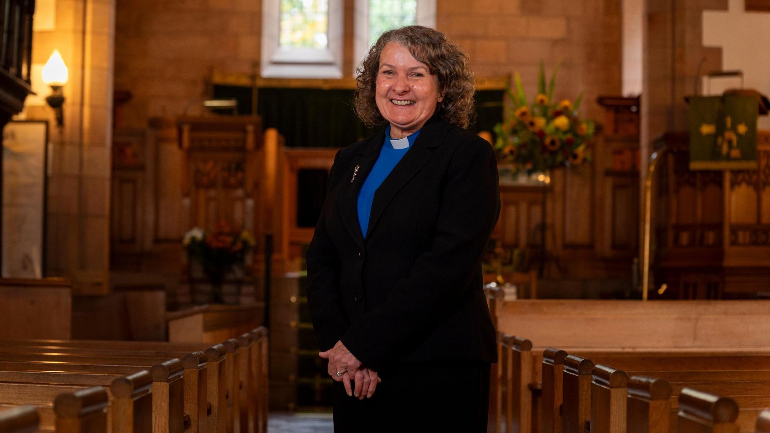 A female minister smiling inside a church with low lighting and flowers and pews in the background