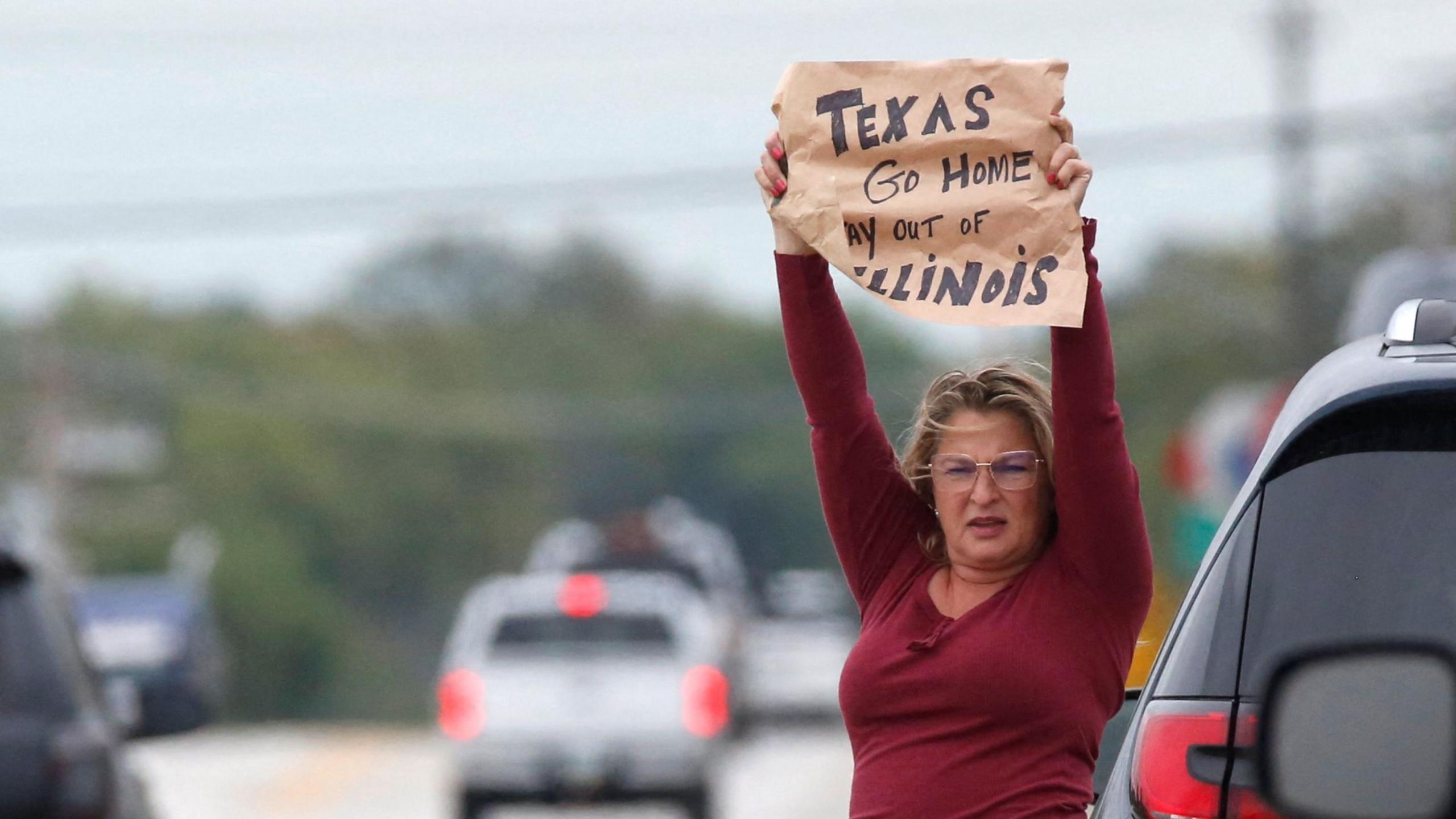 Standing at the side of a road, a woman holds up a sign that reads: "Texas go home, stay out of Illinois"