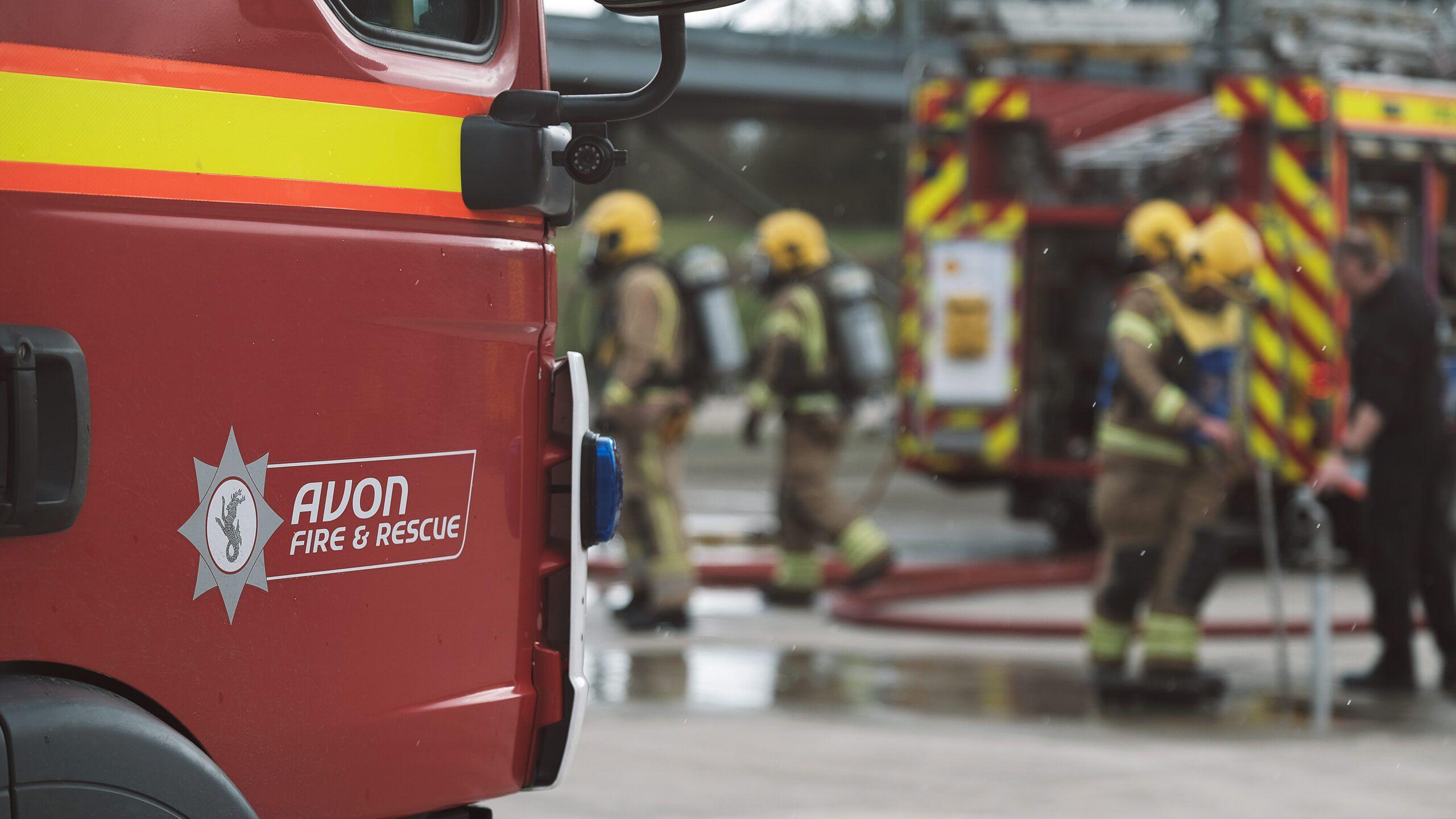 A red fire engine parked in the foreground, with hi-vis decals and the Avon Fire and Rescue branding on the side. In the blurred background there are four firefighters in uniform, pulling a red hose out of the back of another red fire engine.