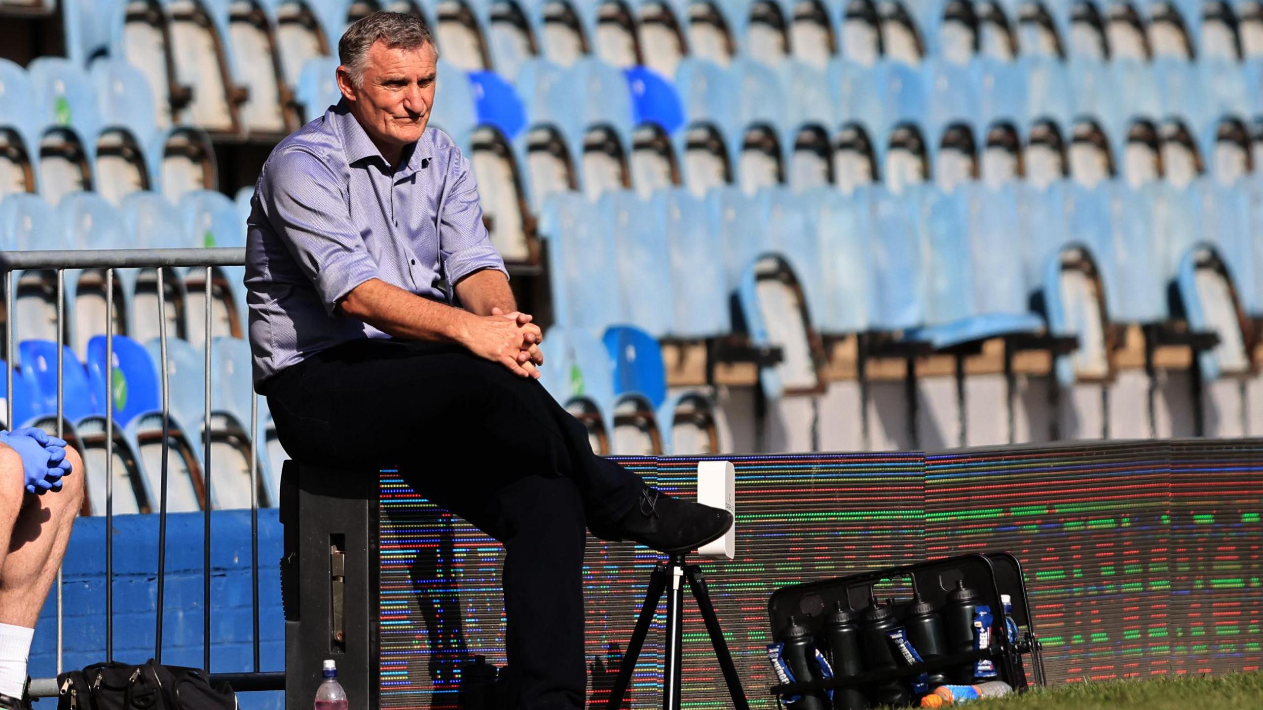 Tony Mowbray, wearing a lilac shirt and black trousers, sits on the advertising boards at London Road Stadium while managing Blackburn Rovers against Peterborough United in 2022