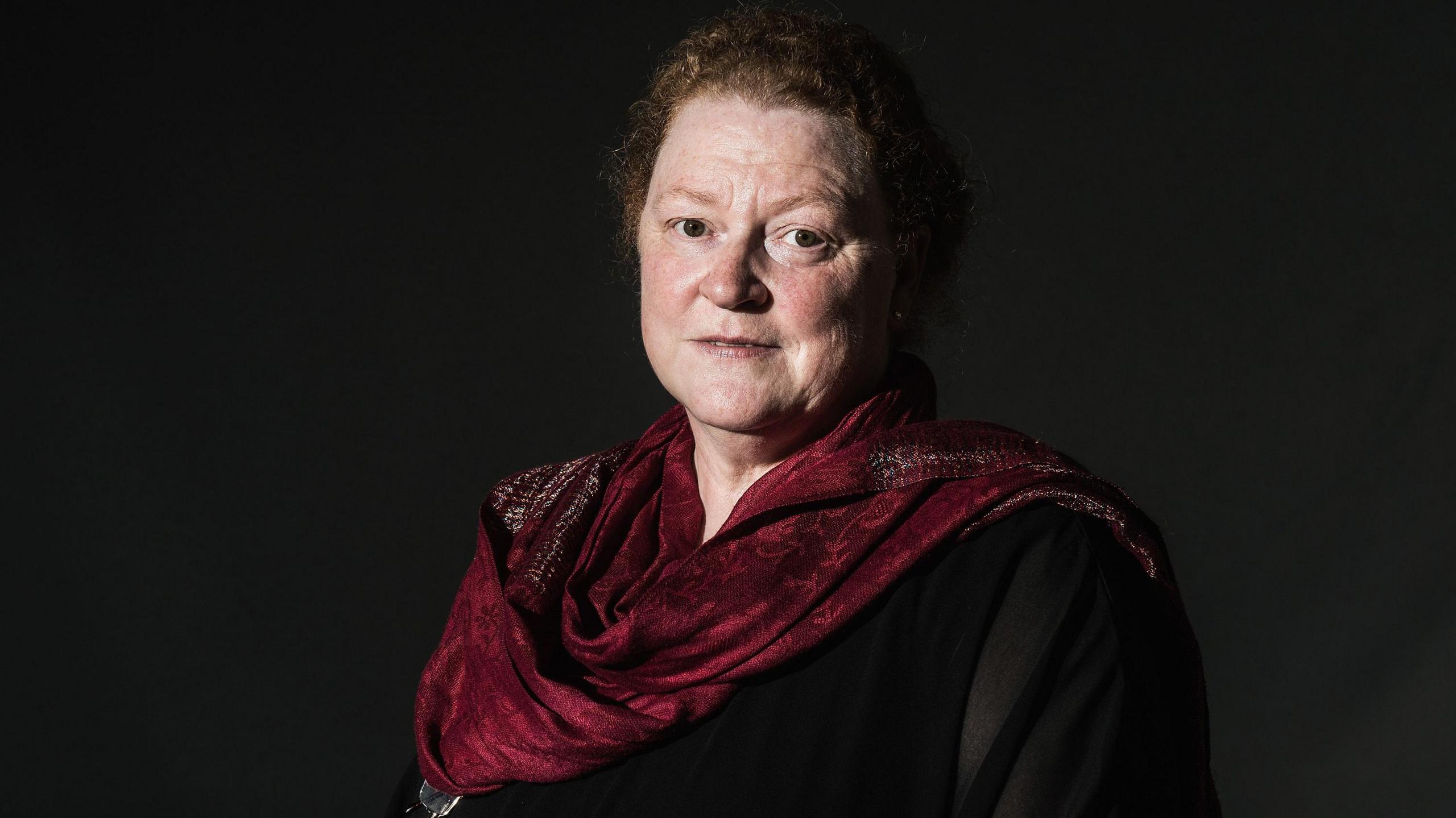 A lady with ginger hair stares at the camera with a serious expression. She is wearing a dark blouse and a dark red scarf.