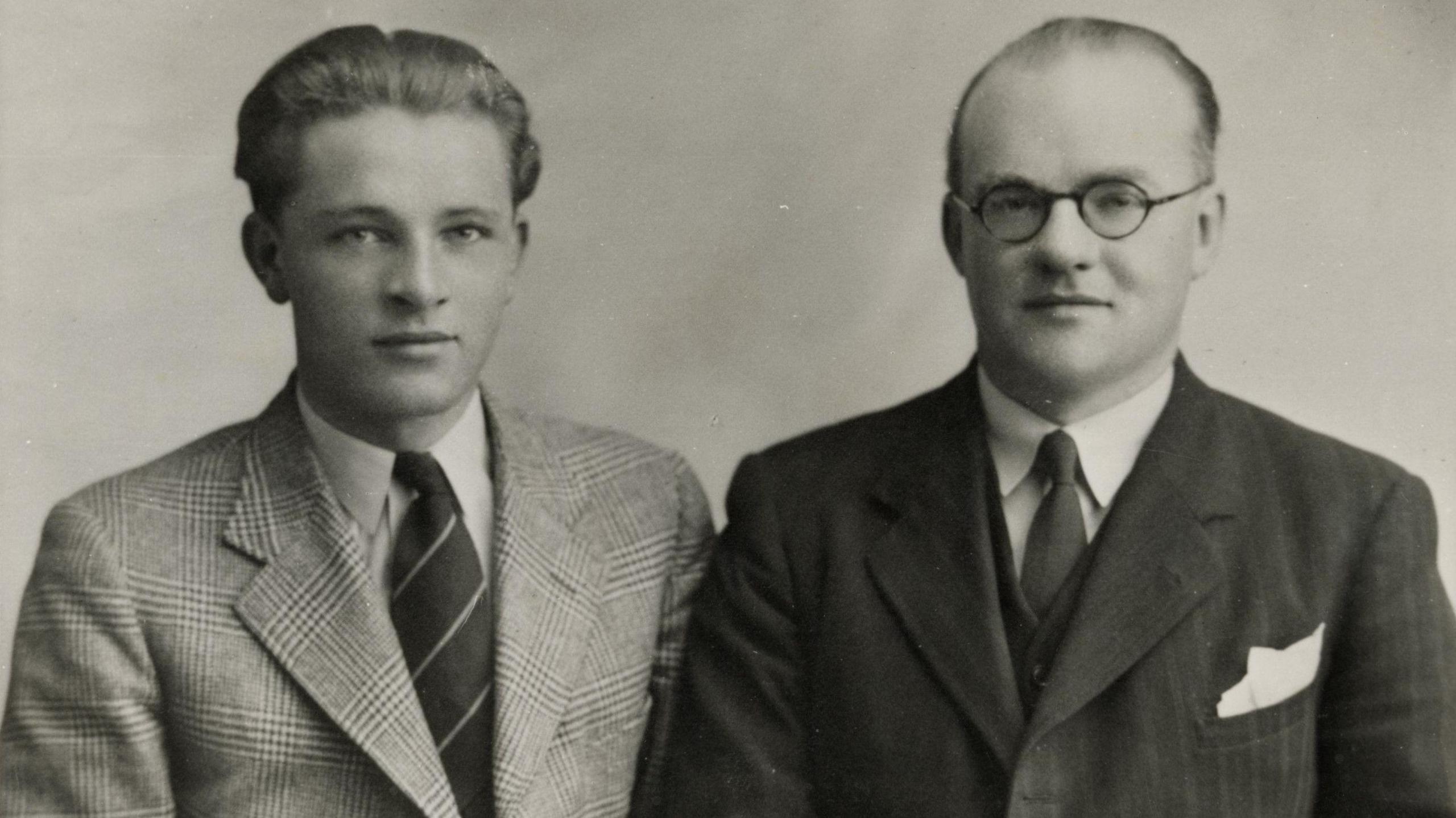 Two men wearing suits in a black and white photograph. The older man is wearing a darker suit with glasses and has a receding hairline.