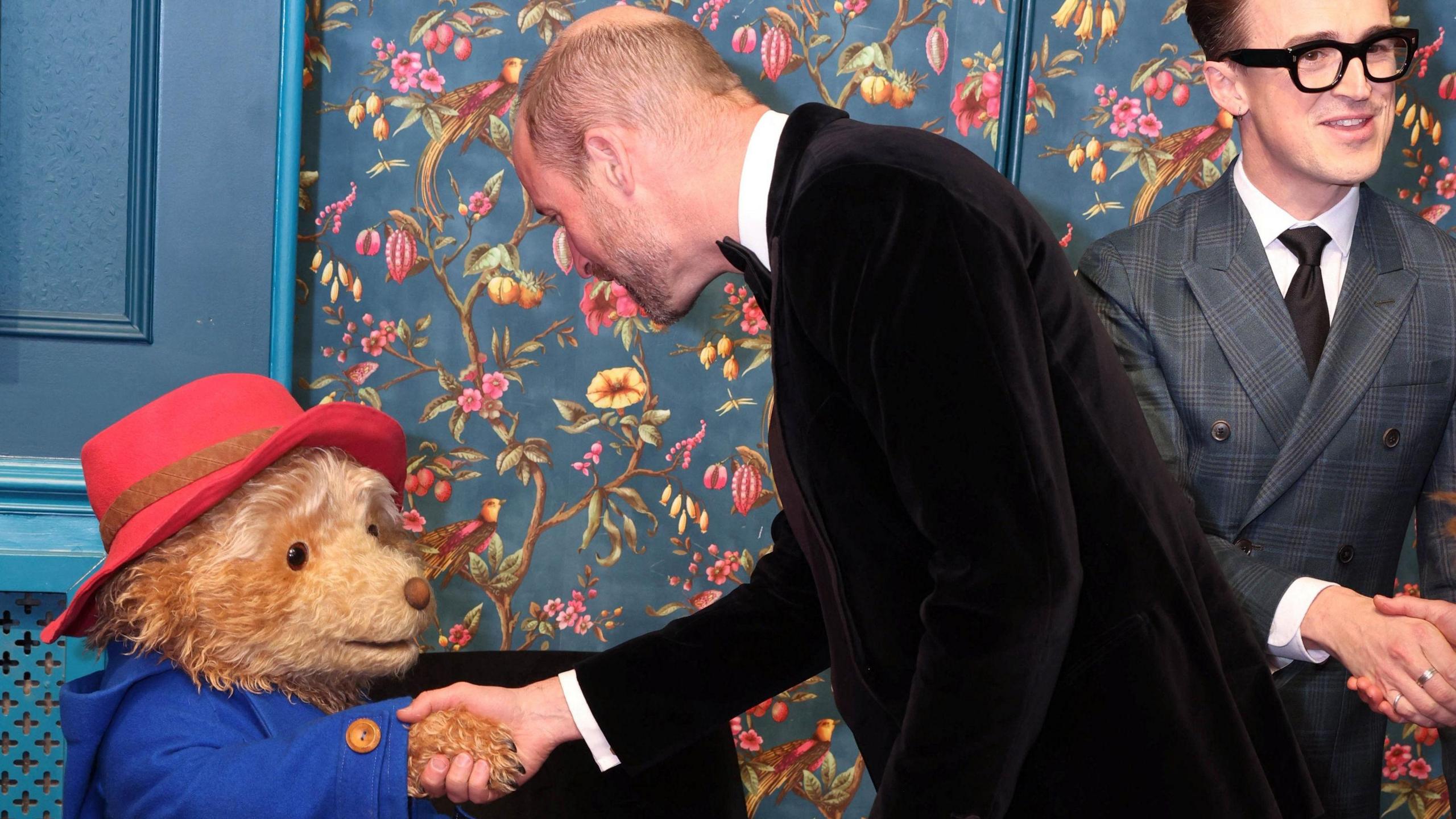 The Prince of Wales, wearing a black tuxedo, shaking the hand of an actor in costume as Paddington Bear - Tom Fletcher from McFly can be seen in the background 