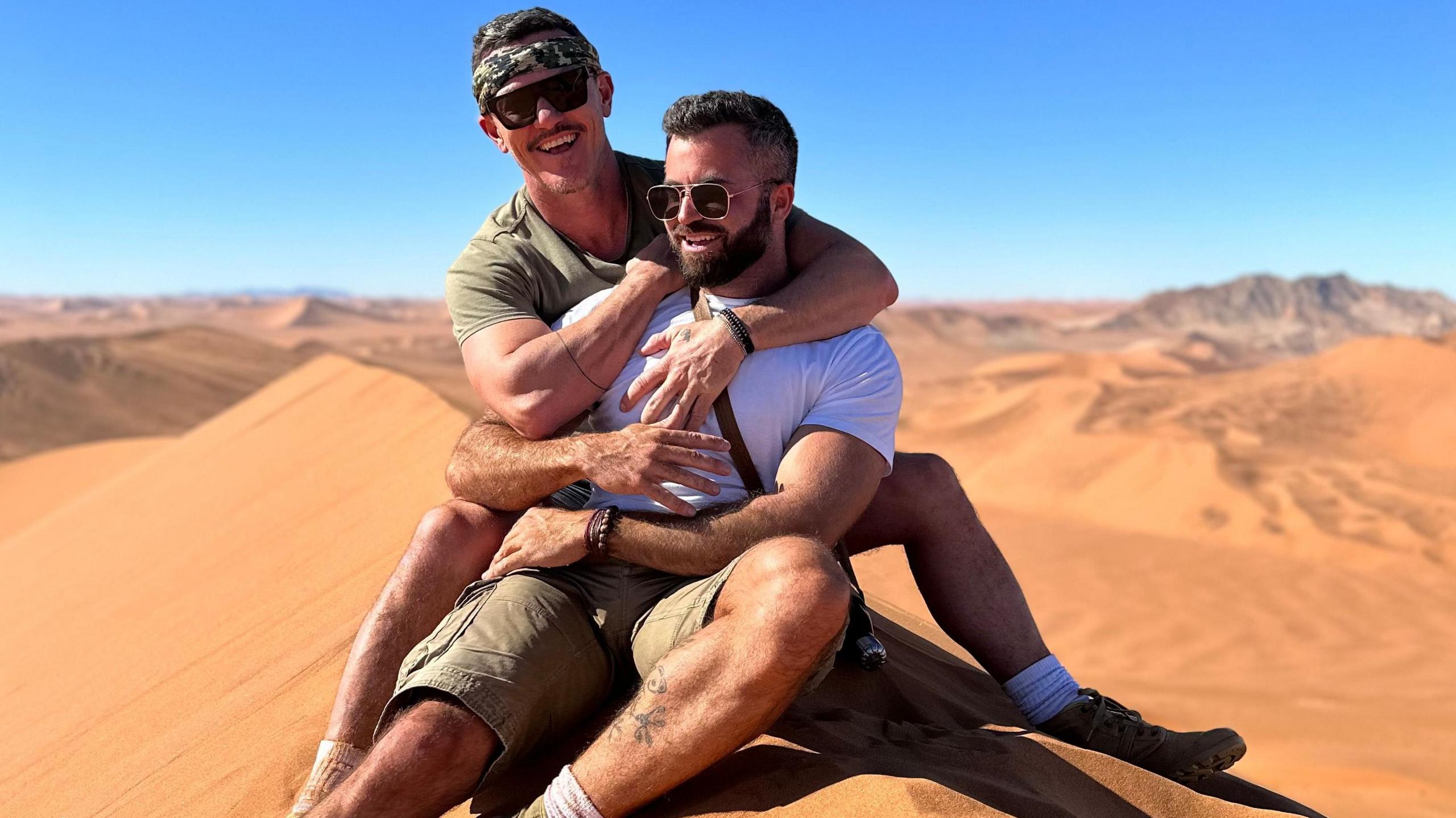 Luke Evans and his partner Fran Tomas cuddling on a sand dune. Luke Evans is behind wearing a khaki green t-shirt and matching headband along with black sunglasses. Fran Tomas is in front wearing a white T-shirt with khaki shorts and boots