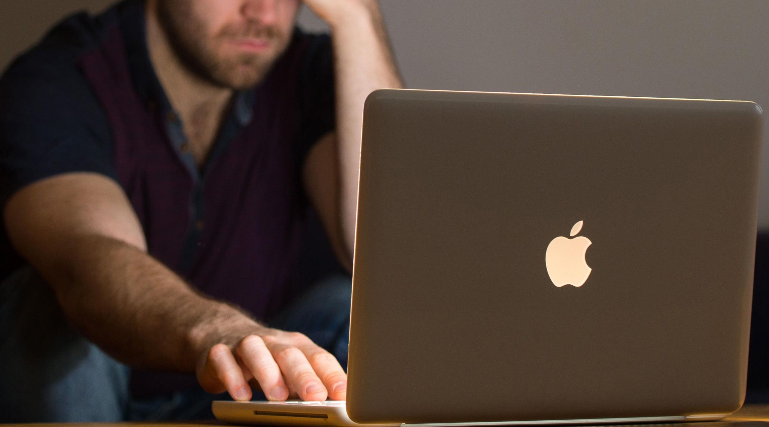 An anonymous man sitting in a dark room with his Apple laptop on a table in front of him. The laptop screen is lighting up his face and he has one hand resting on the mousepad and the other holding his temple. 