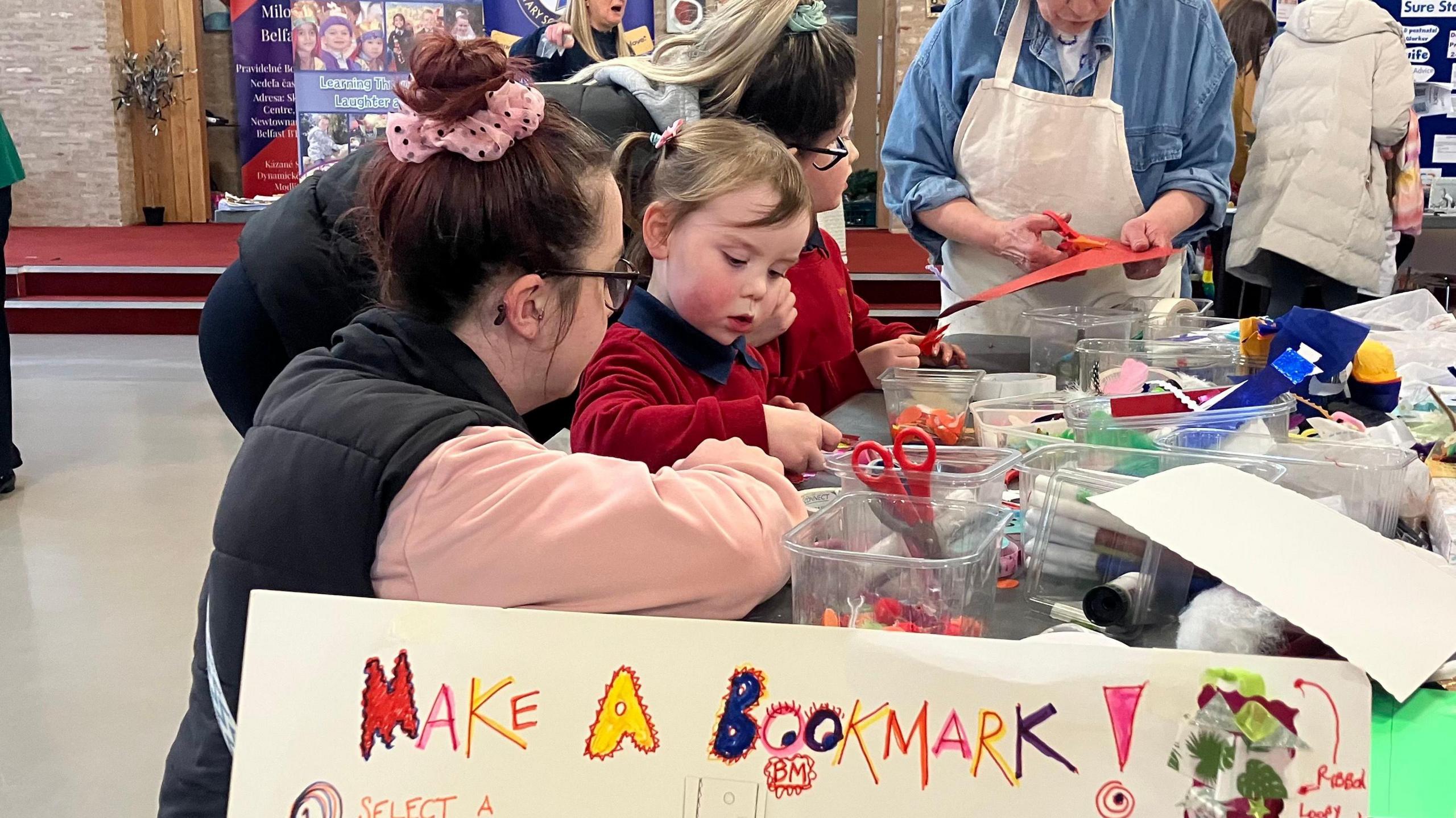 Two young children and two women standing at a table with arts and crafts materials on it. In the foreground a handmade poster that reads: "Make a Bookmark!"