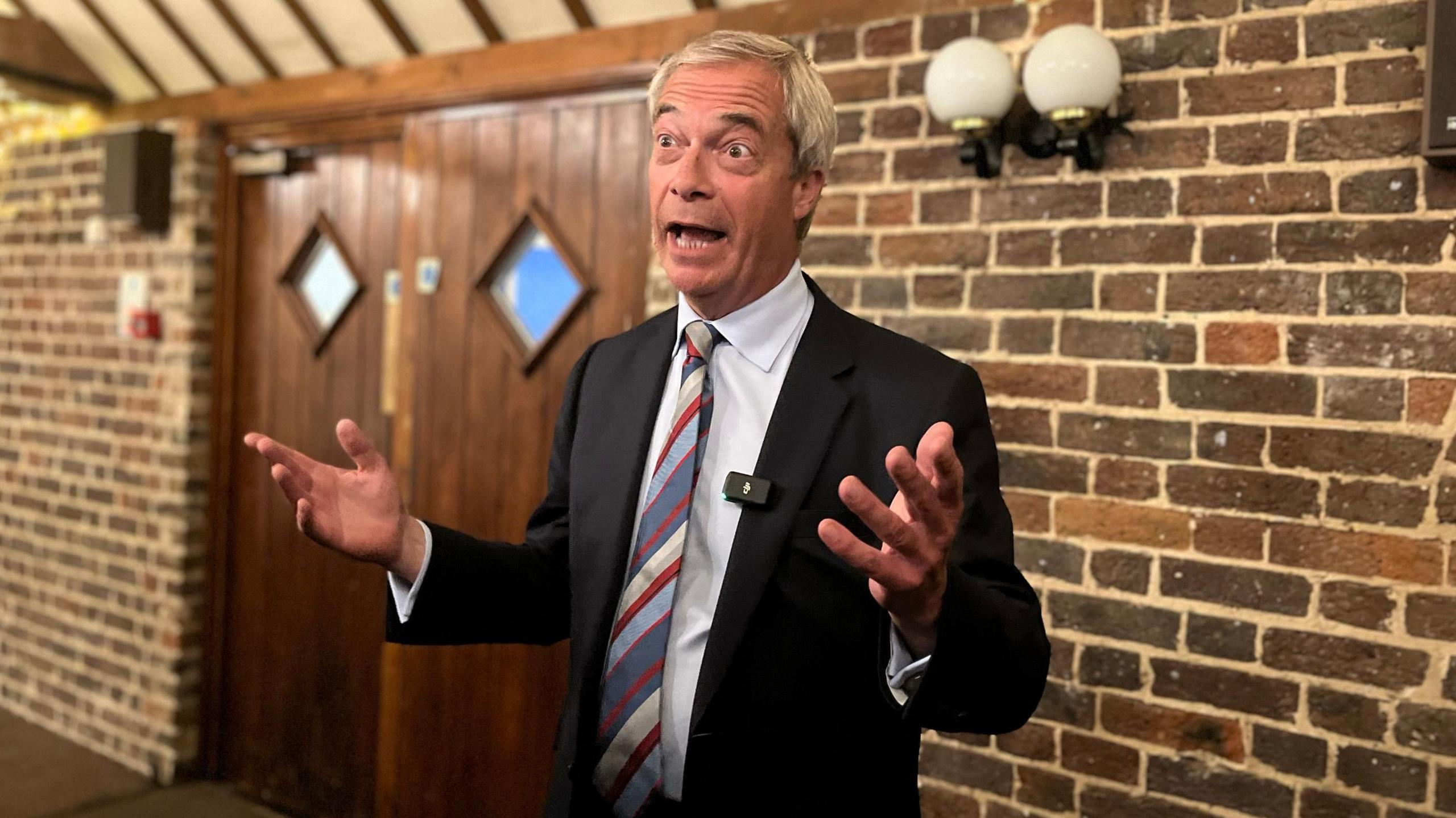 Nigel Farage wearing a white shirt, dark suit and striped tie, stands in front of a brick wall and set of double wooden doors.