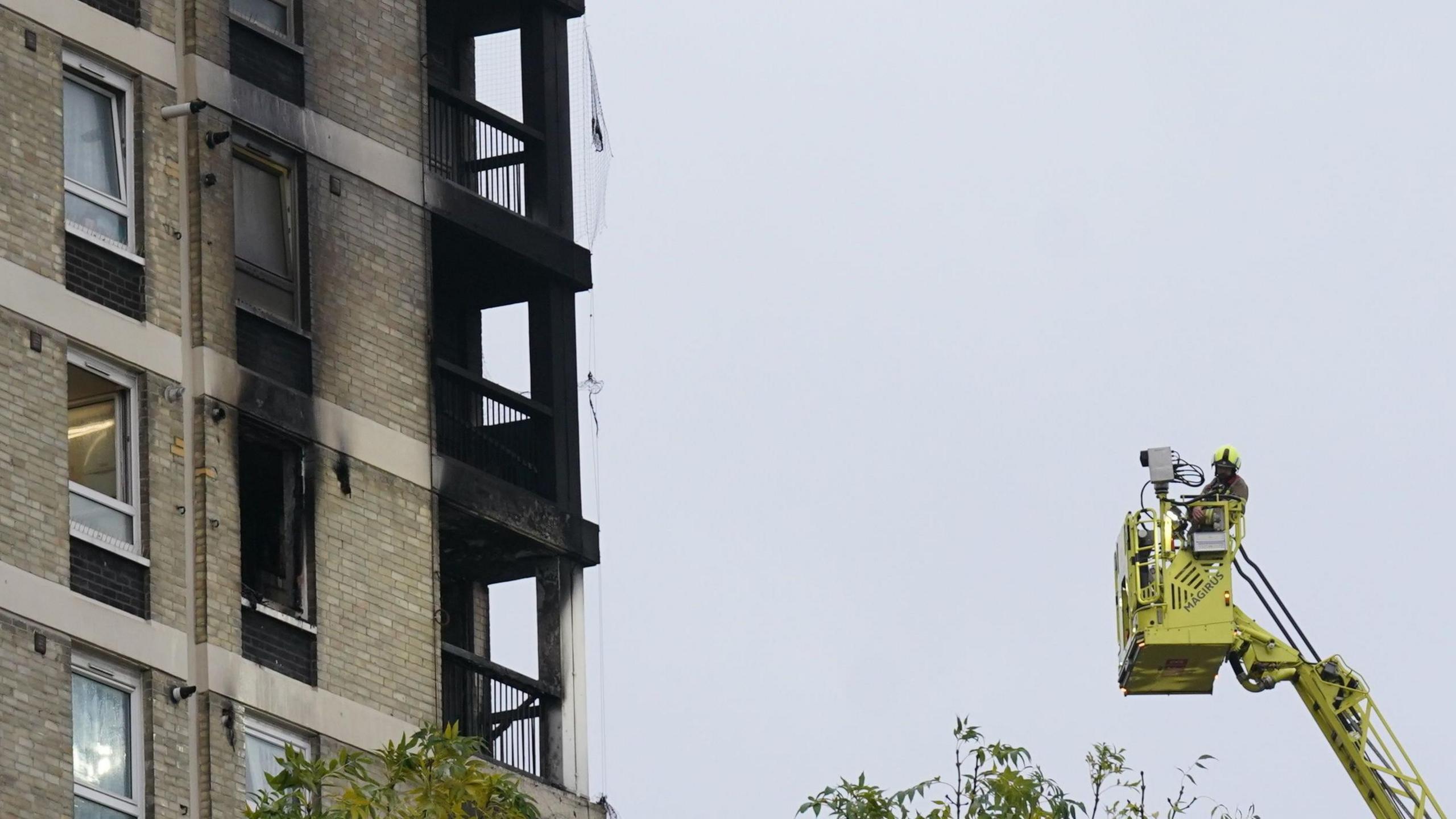 A firefighter at the top of a crane close to the blackened exterior of a tower block. Three stories of balconies have been burnt by the fire and some windows next to the balconies are covered by soot. 