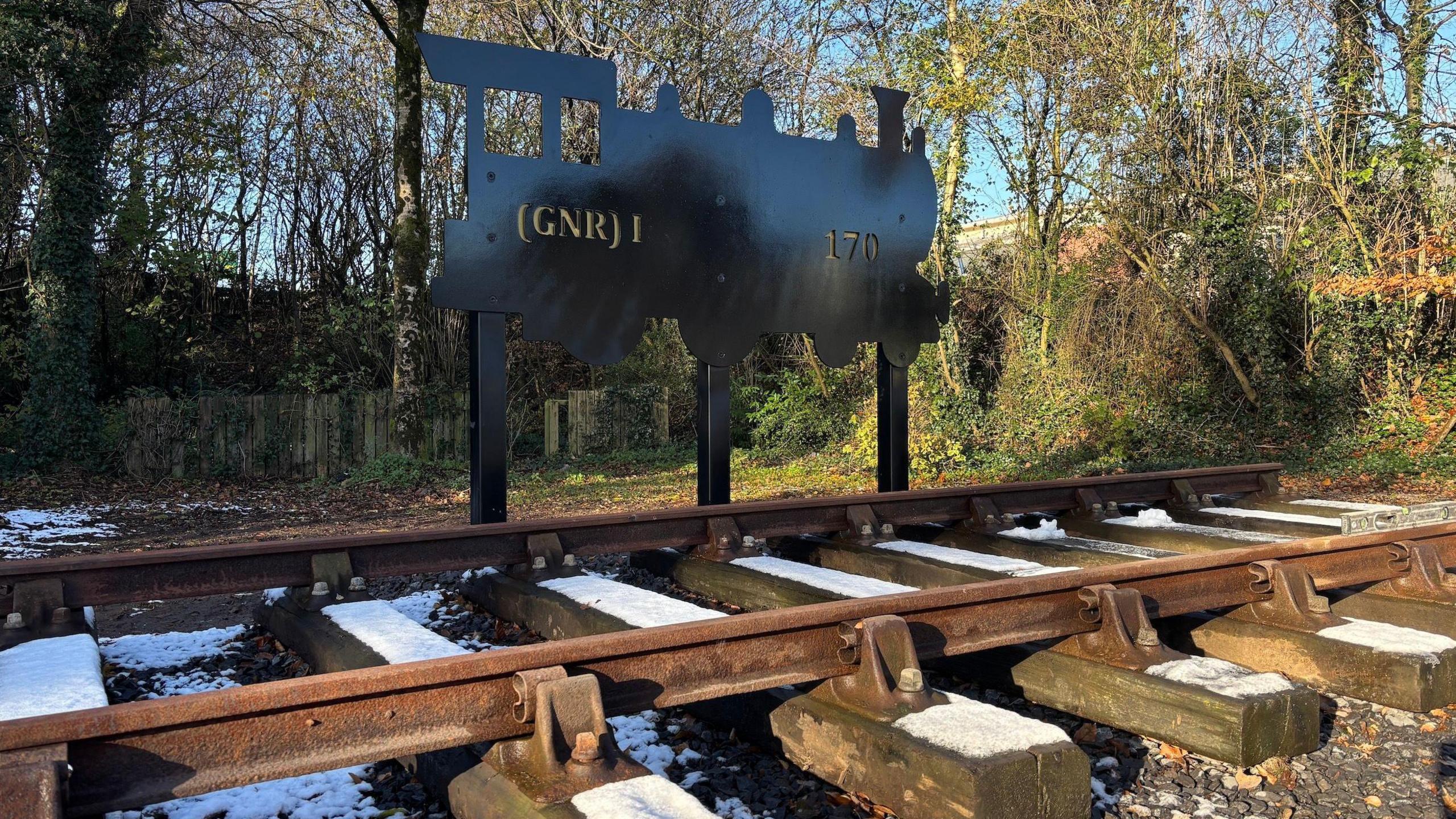 A sculptural silhouette of a train sits alongside a railway track. The depiction of the train bears the mark (GNR) 1 170 in gold lettering. The wooden rail sleepers have a sprinkling of snow upon them. Behind is a fenced garden with trees circling the background.