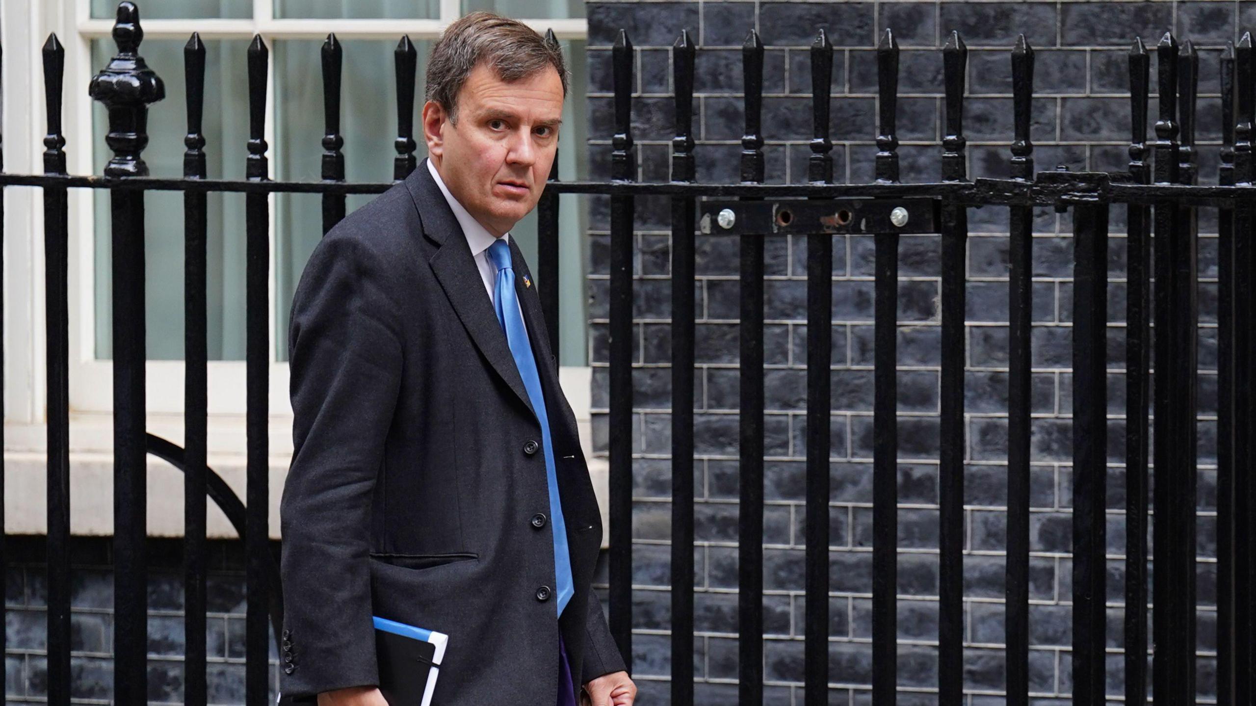 Greg Hands wearing a suit and a blue tie carries a black notebook by the railings in Downing Street