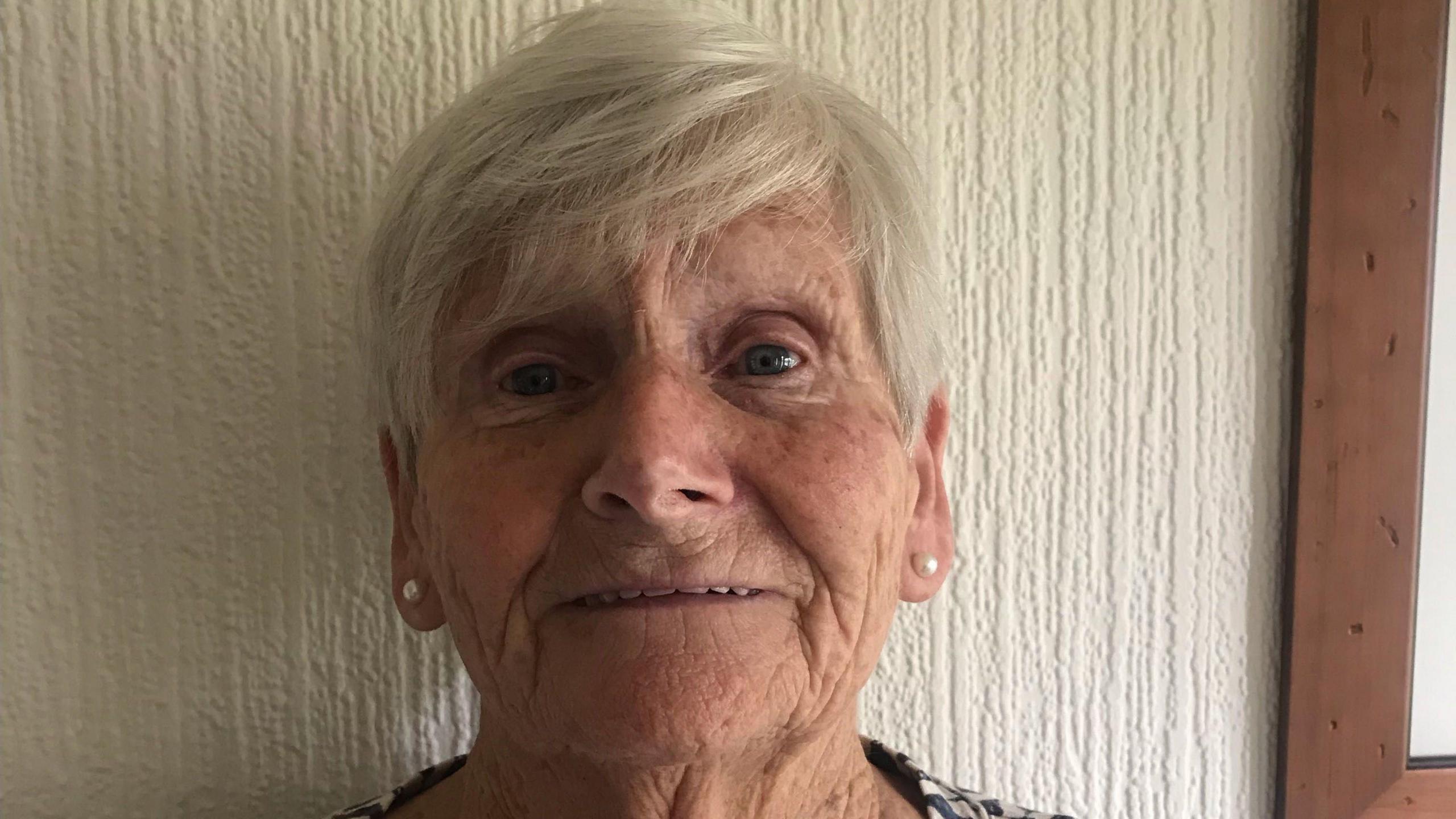 An elderly woman with white short side fringe hair and pearl earrings on against a white wall.