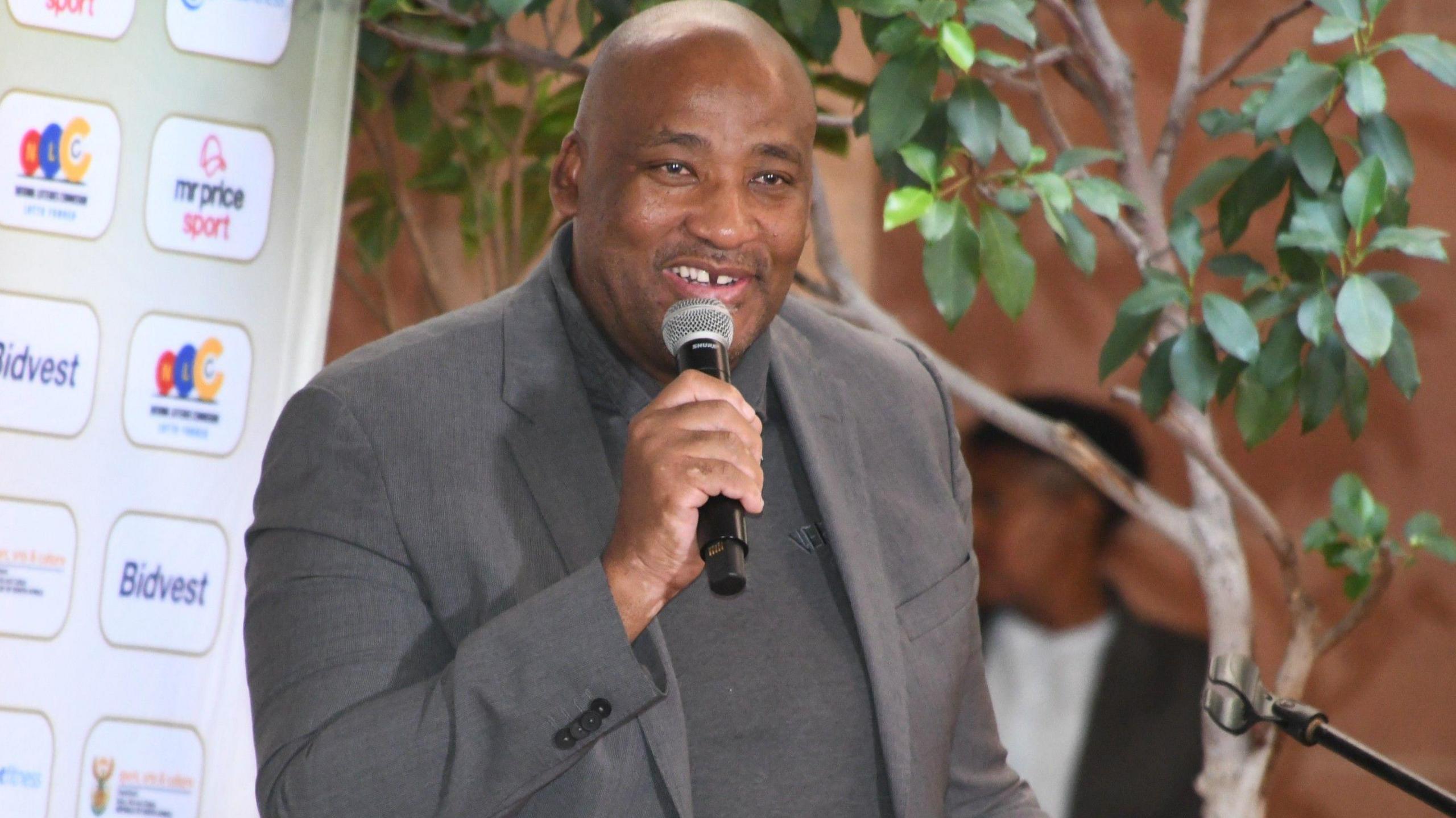 Dressed in a grey suit jacket and grey polo-neck shirt, South African sports minister Gayton McKenzie smiles and holds a microphone to his mouth as he prepares to give a speech with a tree and sponsors board behind him