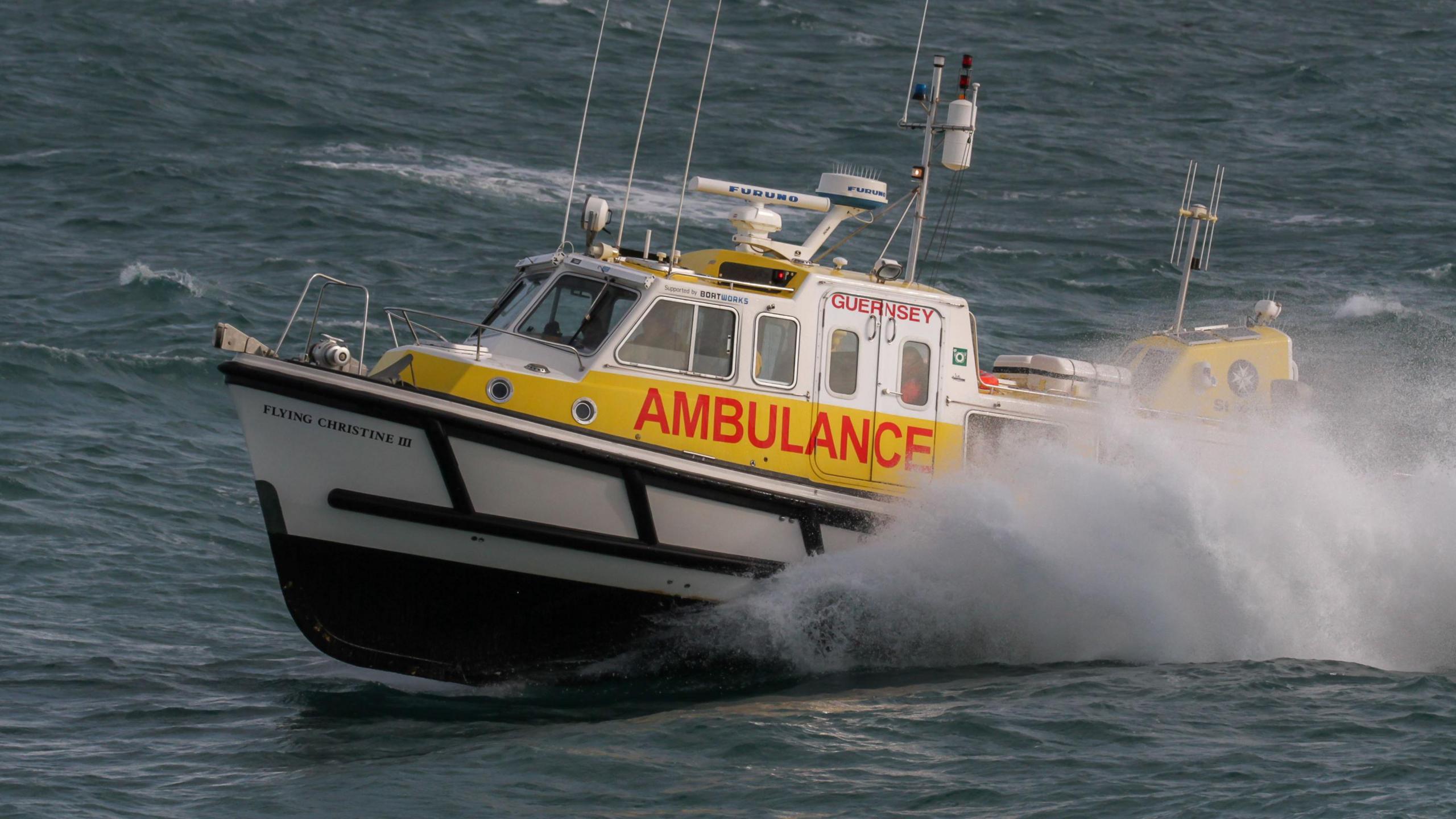 Marine ambulance speeding through the sea. The boat is yellow, white and black. Guernsey and ambulance are written in red and its name Flying Christine III is written in white.