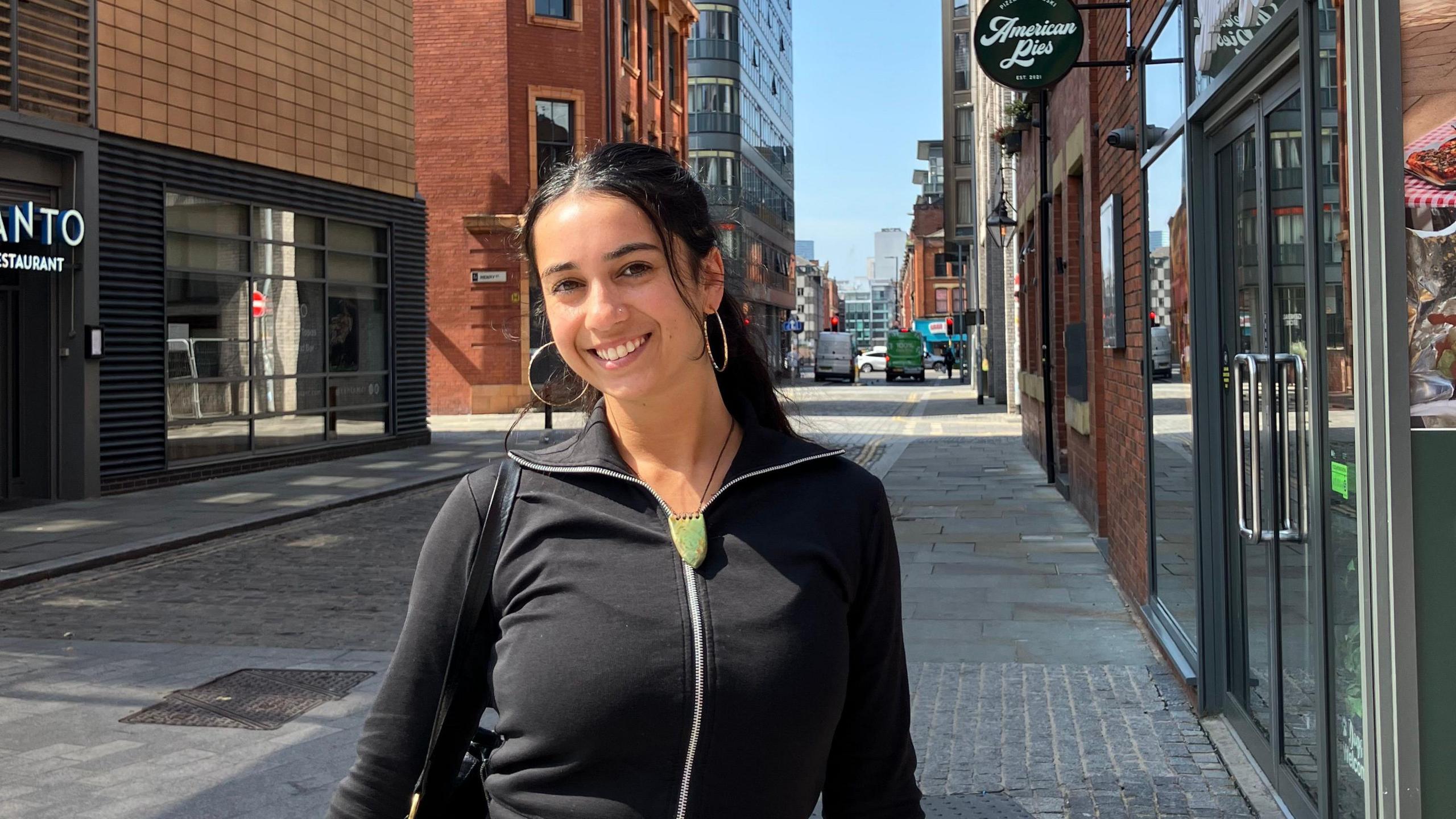 Sufia in the foreground wearing a black zip-up top and light skirt, carrying a black shoulder bag, standing on a paved side street. Behind, a narrow roadway leads to an intersection with traffic lights and a green van. Red-brick and modern buildings line both sides, including a glass-fronted shop with the sign “American Pies”. A large blue-painted utility structure sits to the left. The street has kerbs, paving slabs, and no visible bins, with clear road markings and a few pedestrians in the distance under a bright sky.