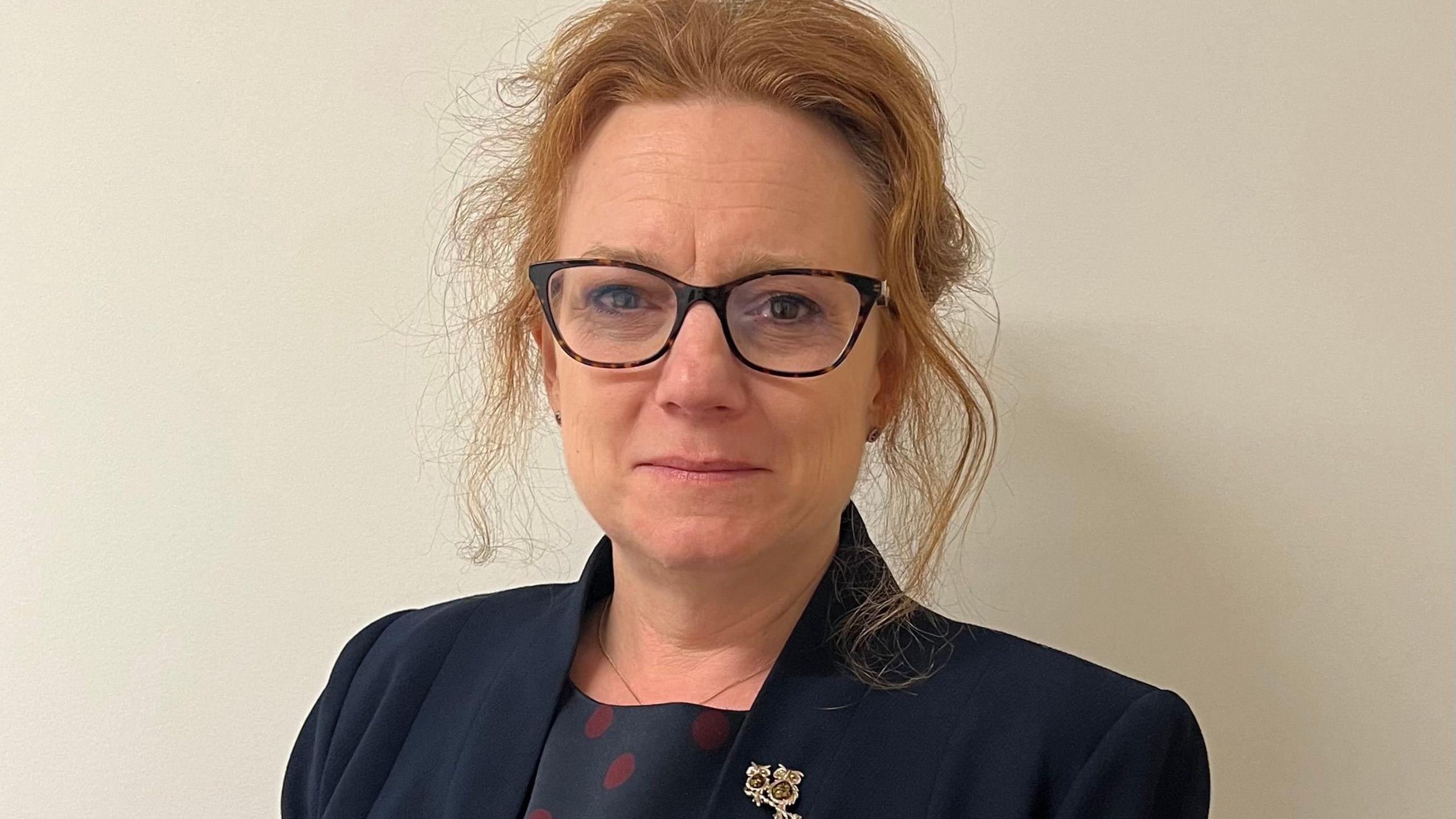 Sarah-Jane Smedmor is wearing a blue suit jacket and top underneath which has dark red spots on it. Her hair is tied back and she is wearing glasses. This is a photo from her shoulders up with a cream background.
