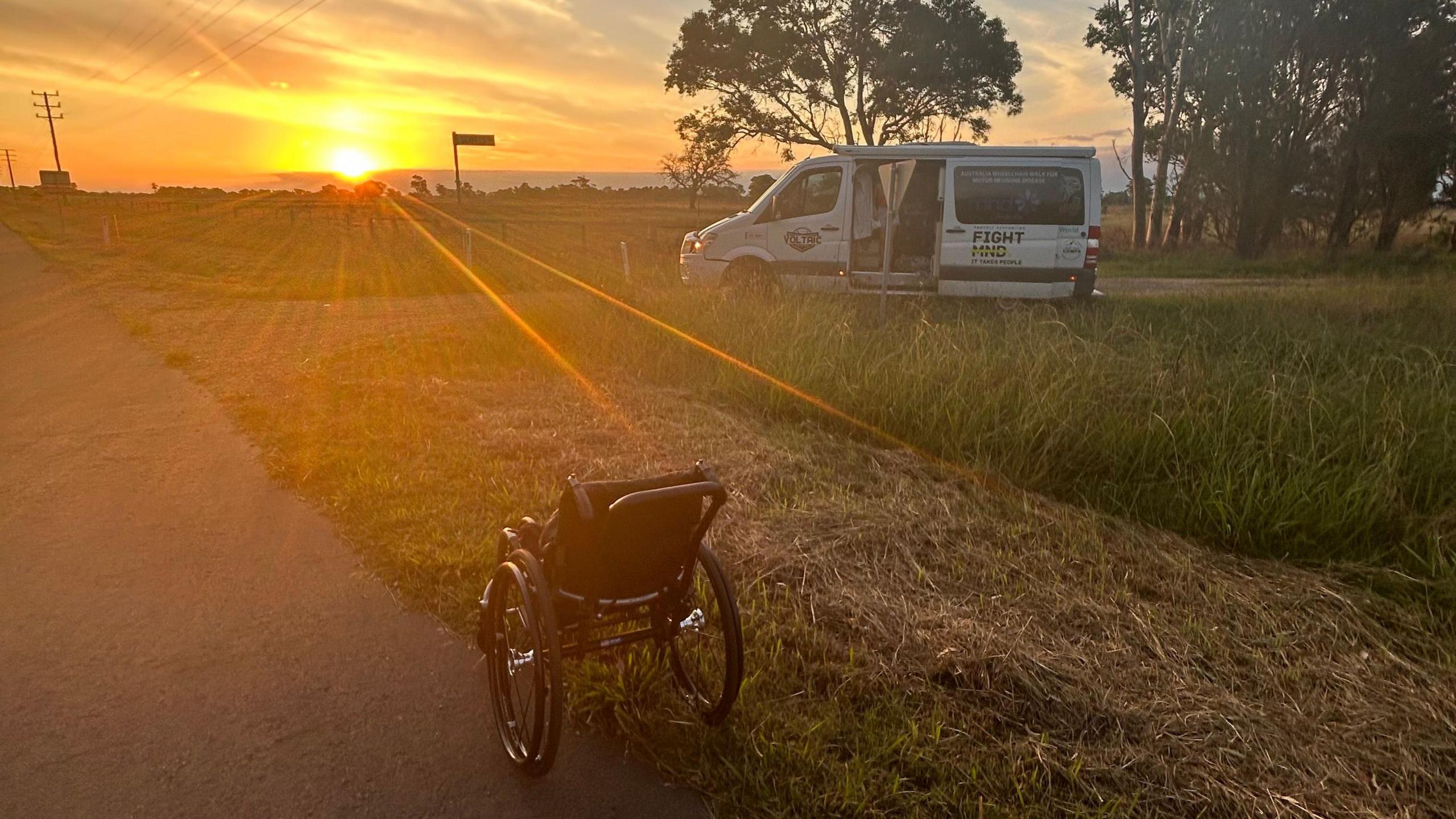 A wheelchair on a road looking out to the horizon where the sun is setting. Billy's van is parked in the background.