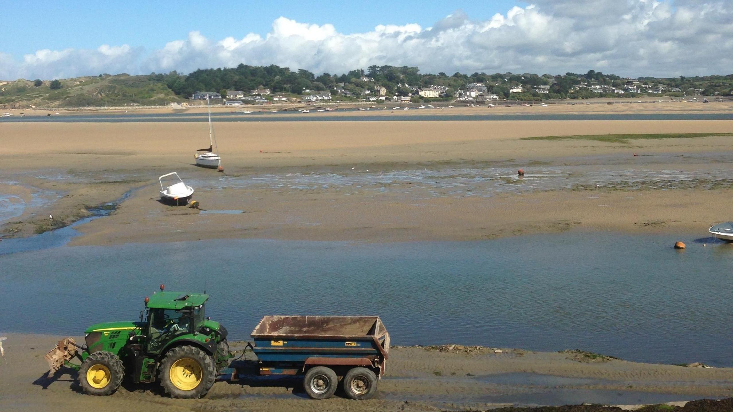 A green tractor pulls a blue trailer with a blue skip on it across the sand by Padstow. The village of Rock can be seen in the background across the bay. Three boats are on a sand bank.
