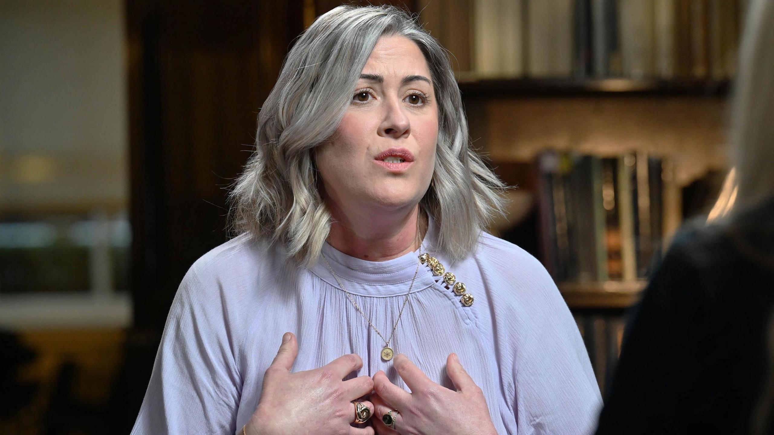 Caroline Darian gesticulates in a picture taken during her interview, bringing her hands up to her chest. She is a woman in her 40s with her hair in a bob, wearing gold jewellery.