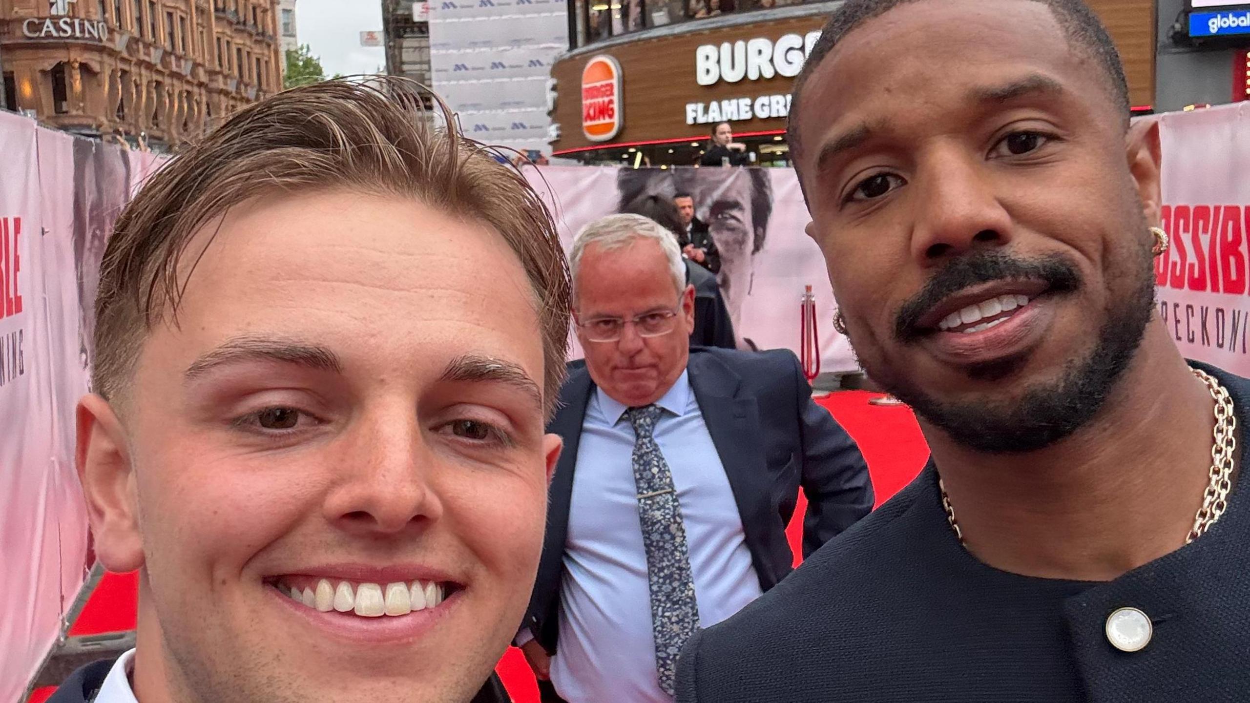 A smiling Wilf Elliott posing for a photo with the actor Michael B. Jordan. They are standing on a red carpet at a film premiere and a man wearing a navy suit is standing behind them.