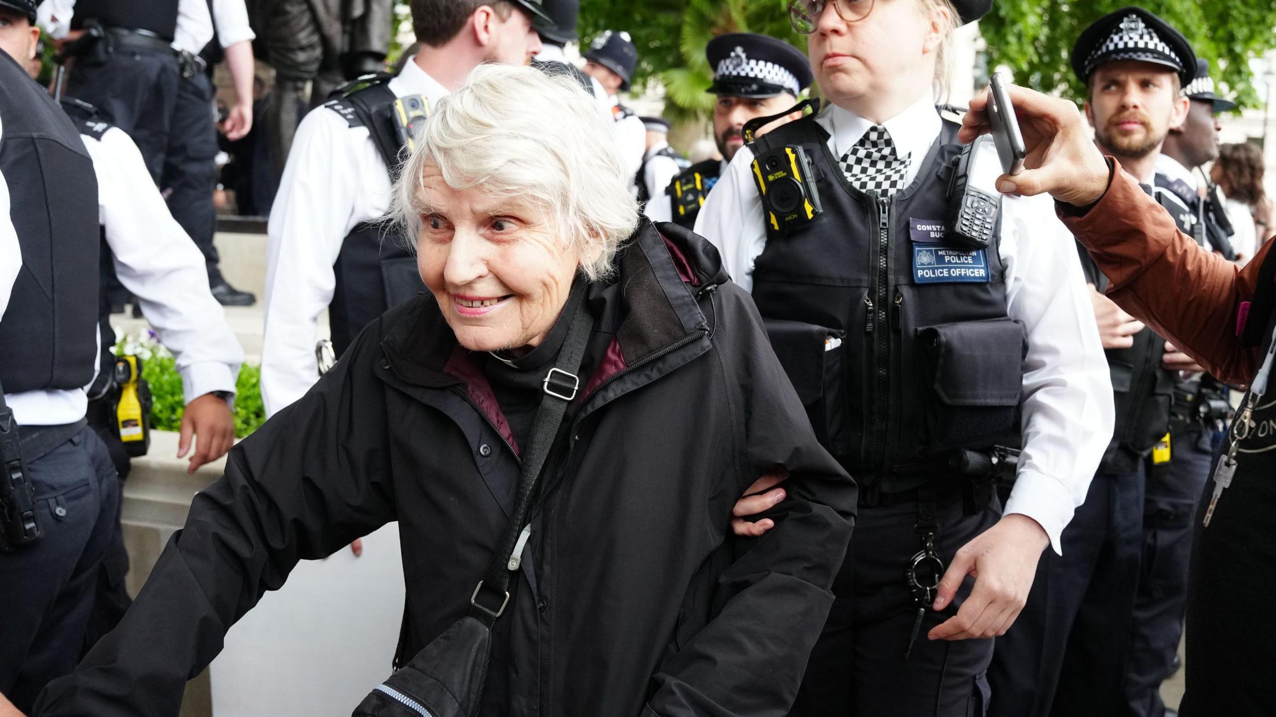 Reverend Sue Parfitt with two police officers either side of her. She is wearing a vicar's collar and a back coat while sat near white stones.