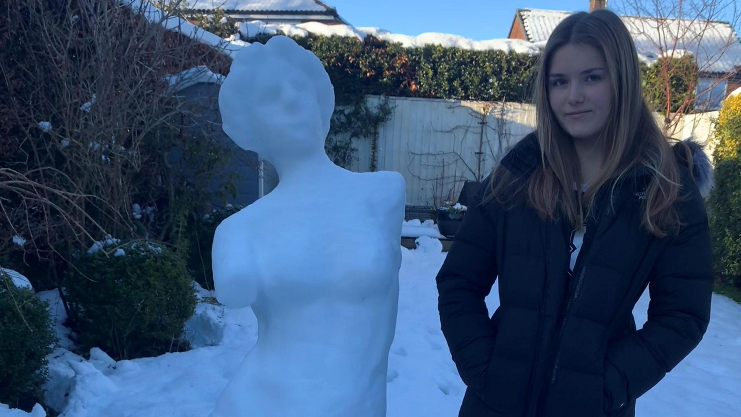 A snow sculpture of the Venus de Milo and a teenage girl with long blonde hair in a black duffel coat.