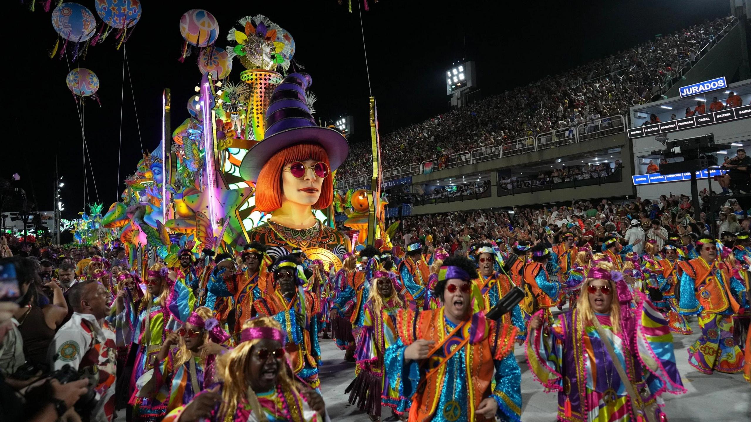 A colorful parade in Rio de Janeiro in Brazil with people wearing bright, colourful costumes and head dresses. There's a figure of a head with a witch hat on a float and the parade is being watched by a large crowd in a busy stadium