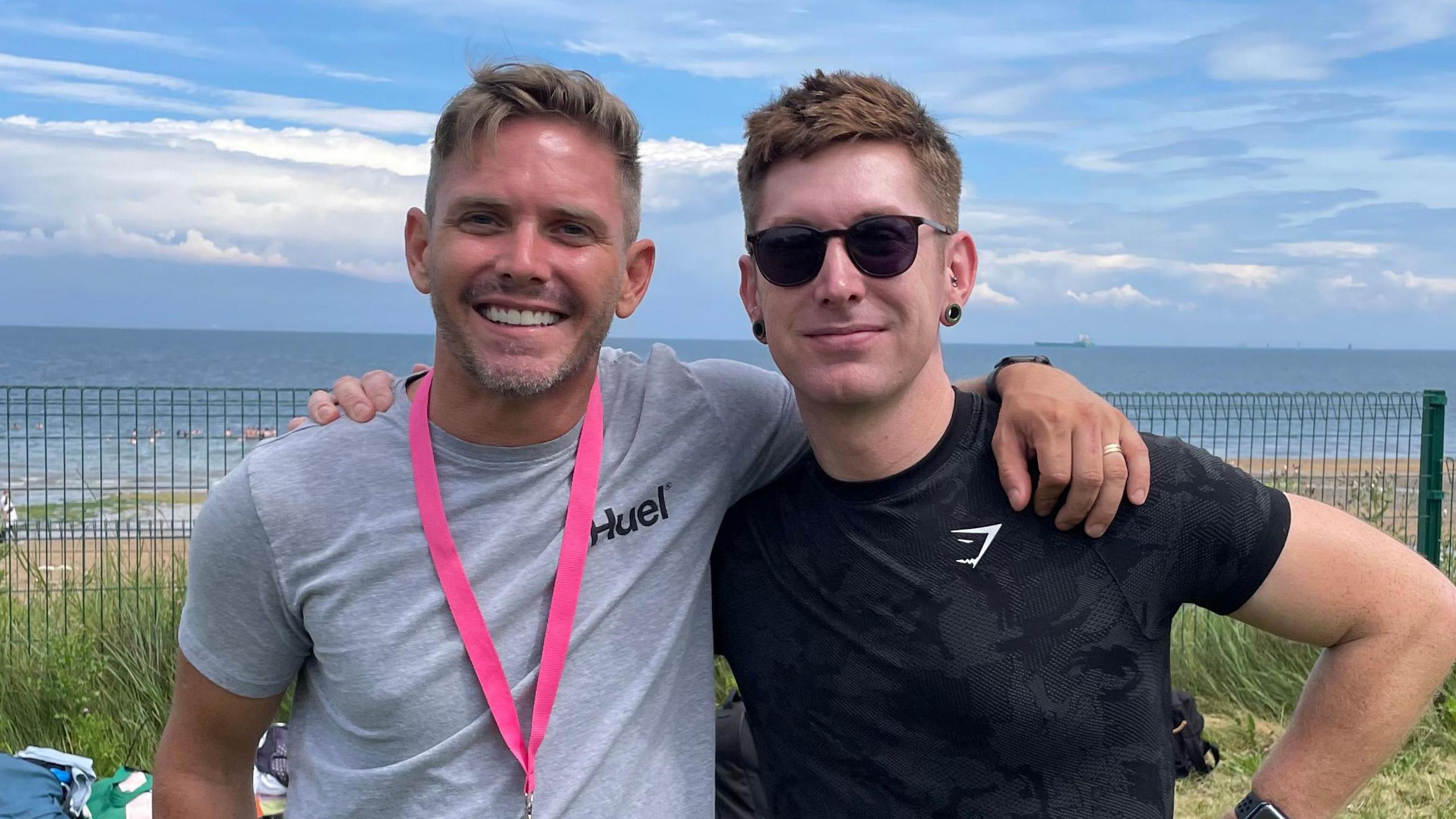 Two men, standing with their arms around each other's shoulders. The man on the left has short brown hair and he is wearing a grey t-shirt and a pink lanyard. The man on the right has short brown hair and he is wearing a black t-shirt and sunglasses. They are standing on a patch of grass and the beach and see is visible behind them. 
