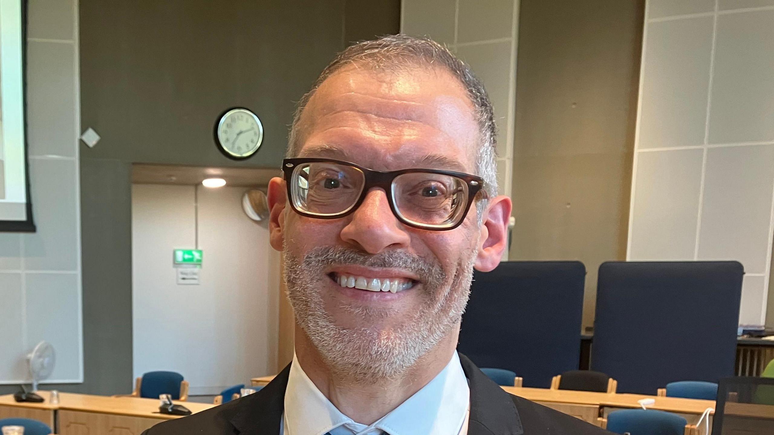 Adam Zerny smiles at the camera. He has short grey hair and a beard. He wears black glasses, a black suit, white shirt and blue tie. He stands within a council chamber.