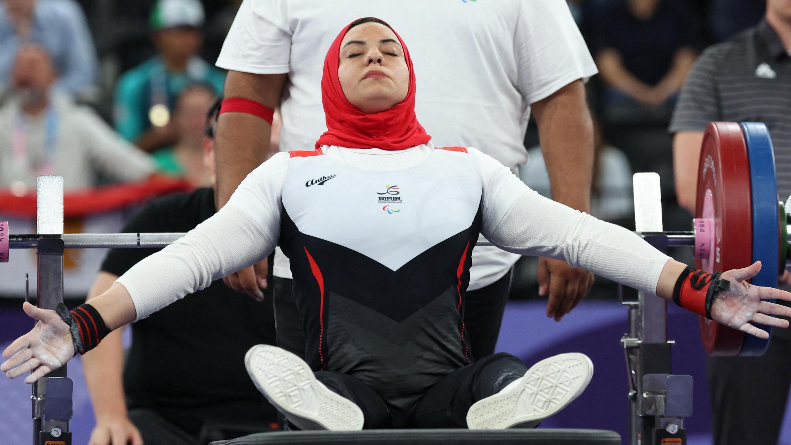 Rehab Ahmed, wearing a red headscarf and a white and black tracksuit, closes her eyes and spreads her arms out wide in a moment of focus before competing in a para-powerlifting event