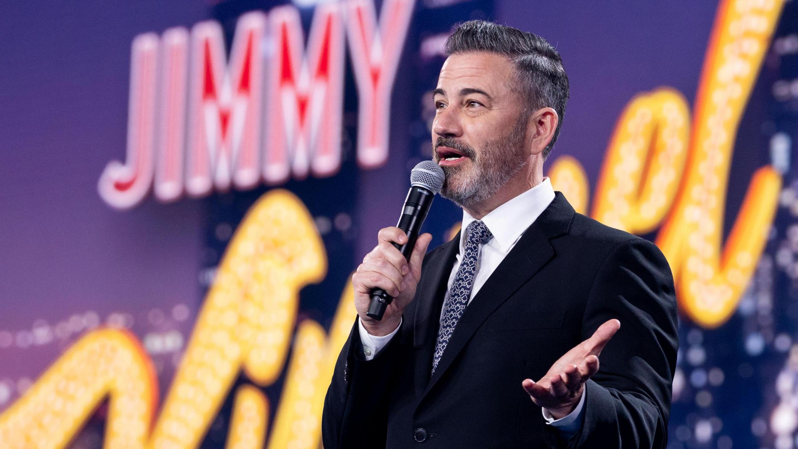 Jimmy Kimmel speaking into a microphone in front of a giant sign bearing his name in May 2025
