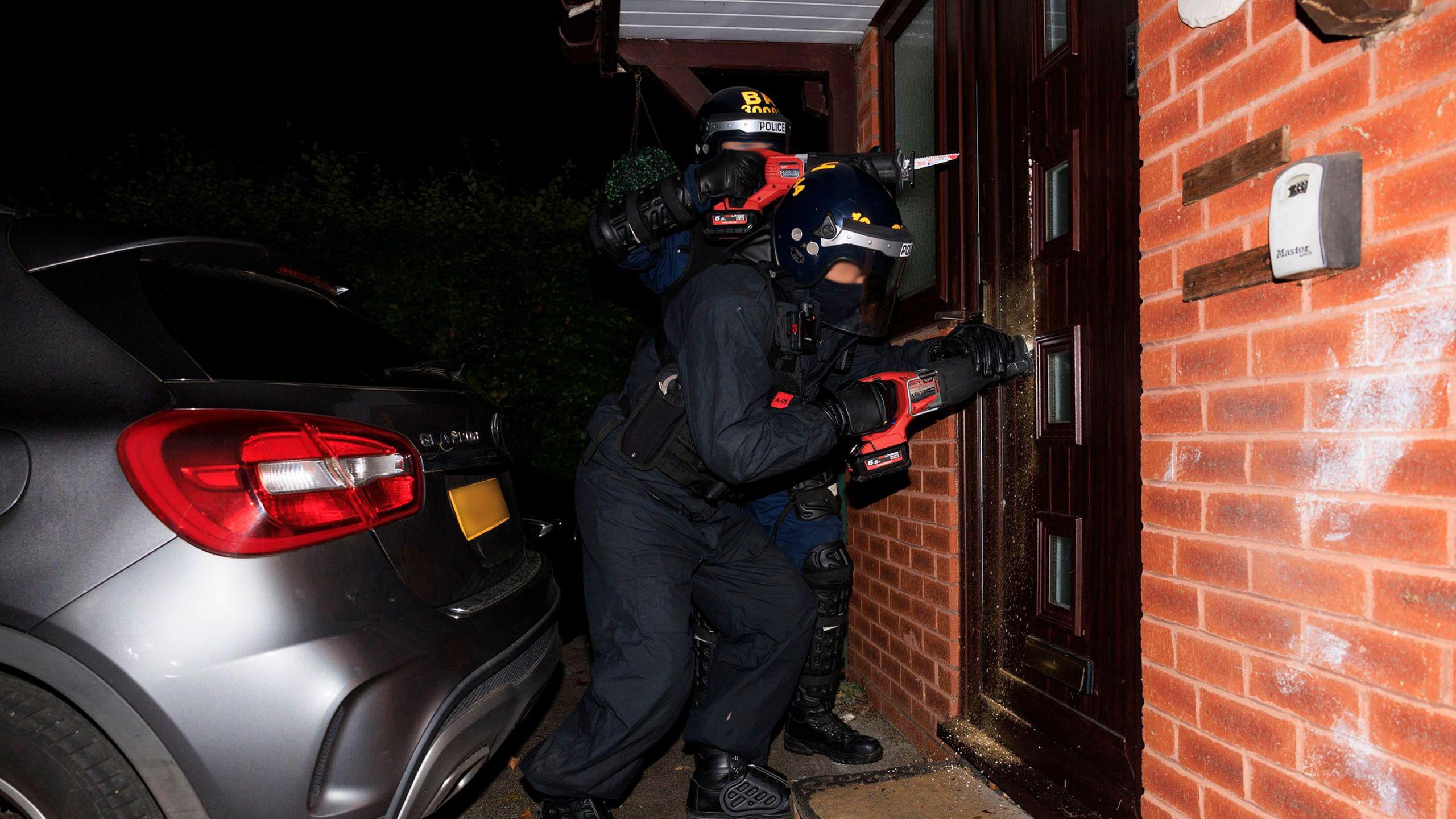 Two police offices holding electric screwdrivers try to get  into a front door of a property