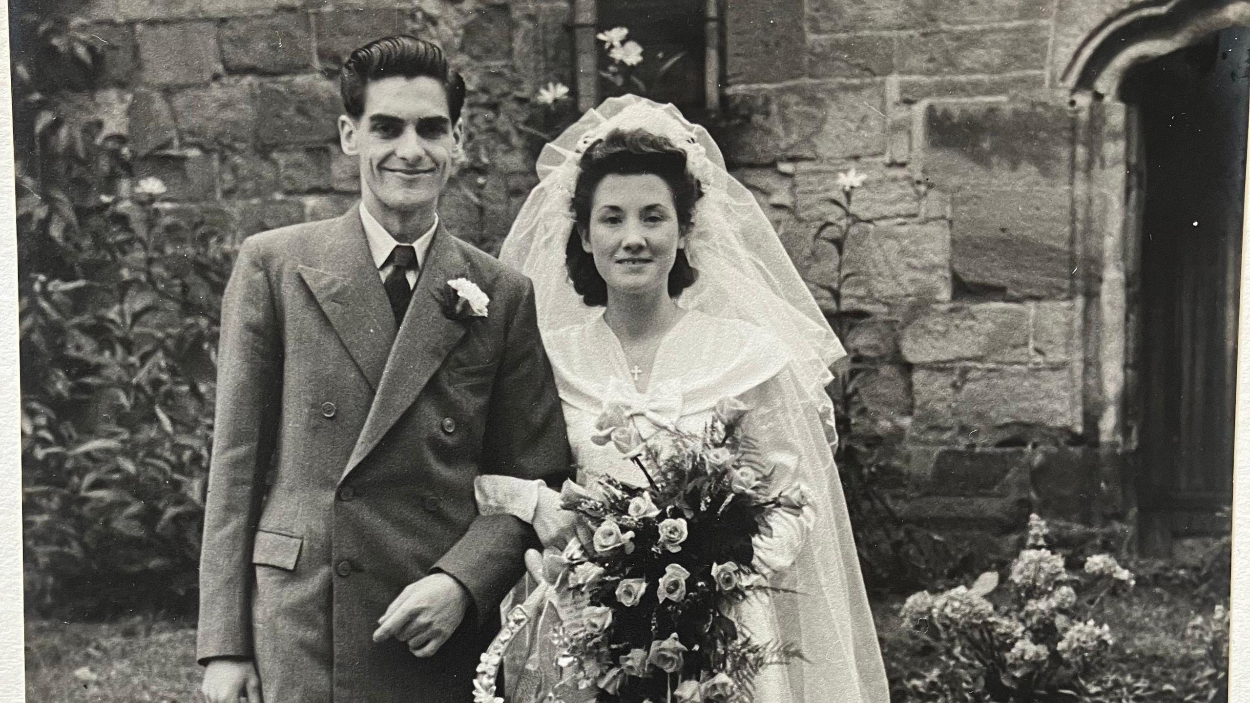 A black and white photo of a man in a dark suit with black slicked back hair with a woman in a wedding dress holding his arm with a bouquet of flowers. 