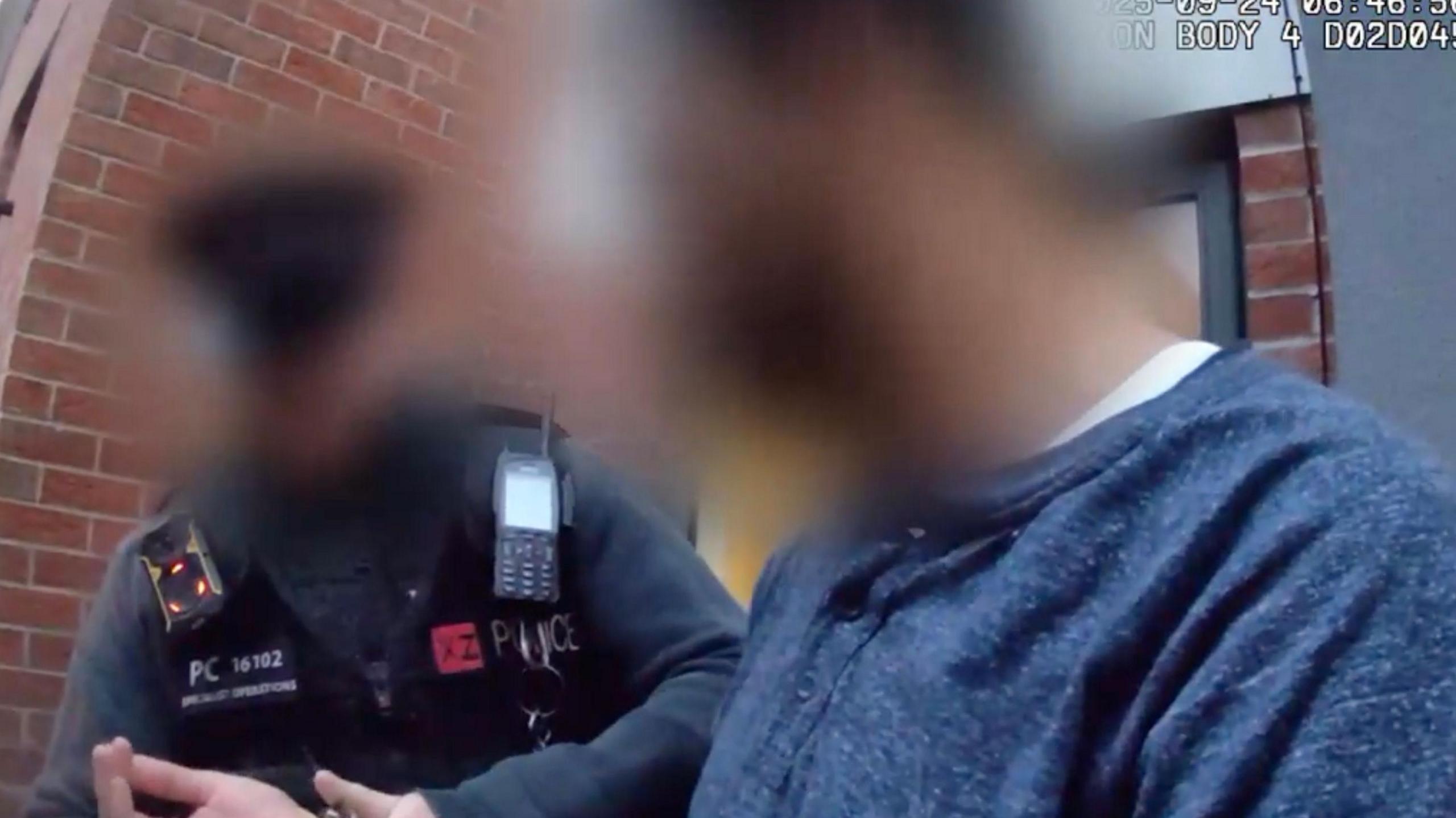 A screenshot from a police body camera of a man being handcuffed by a police officer in a grey uniform. Both the man and the police officer's faces have been blurred out. 