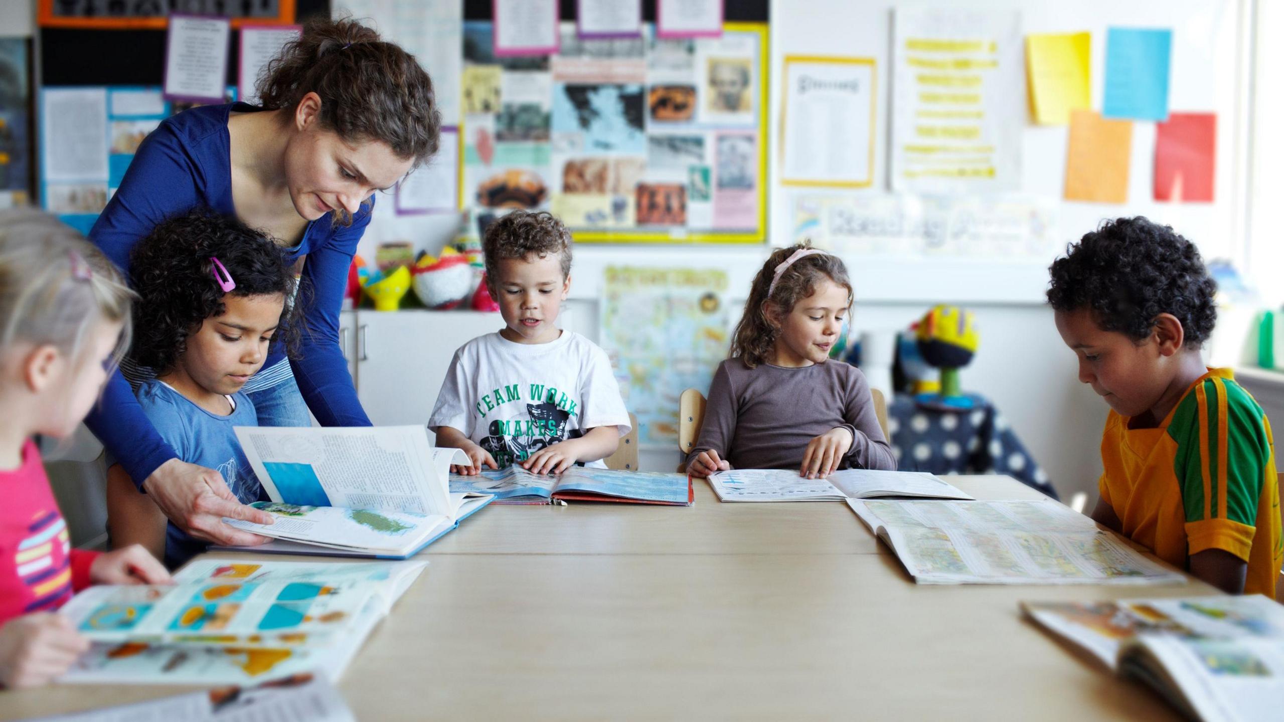 Five children with text books sit around desks in a school classroom while a teacher helps one of the children with her book.