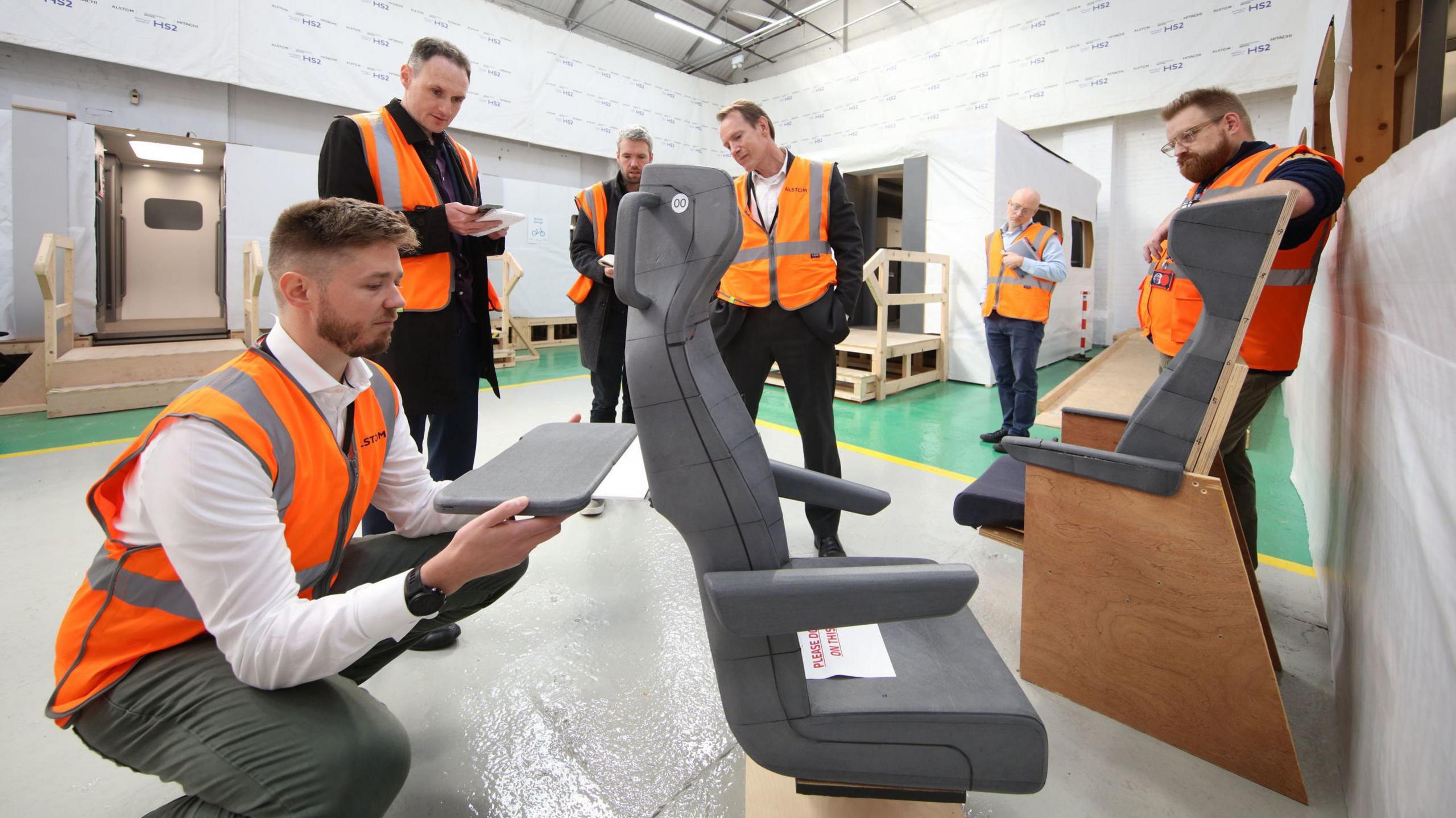 Six men in orange hi-vis inspect a mock seat on the factory floor in Derby