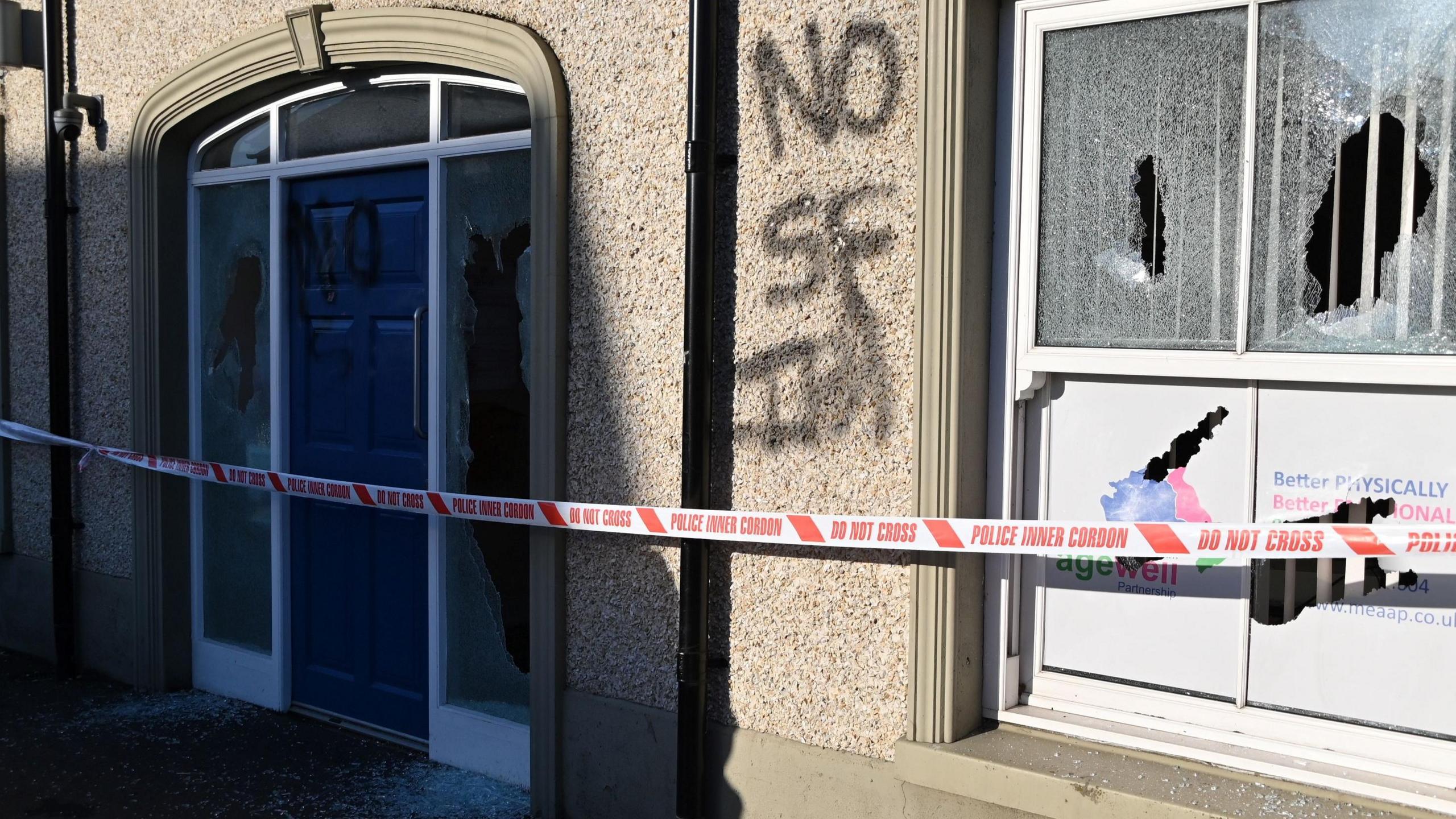 The front of a grey pebble-dashed building with several of it's windows smashed and black graffiti that reads "No SF IRA" sprayed on the walls. There is police tape across the front of the building.