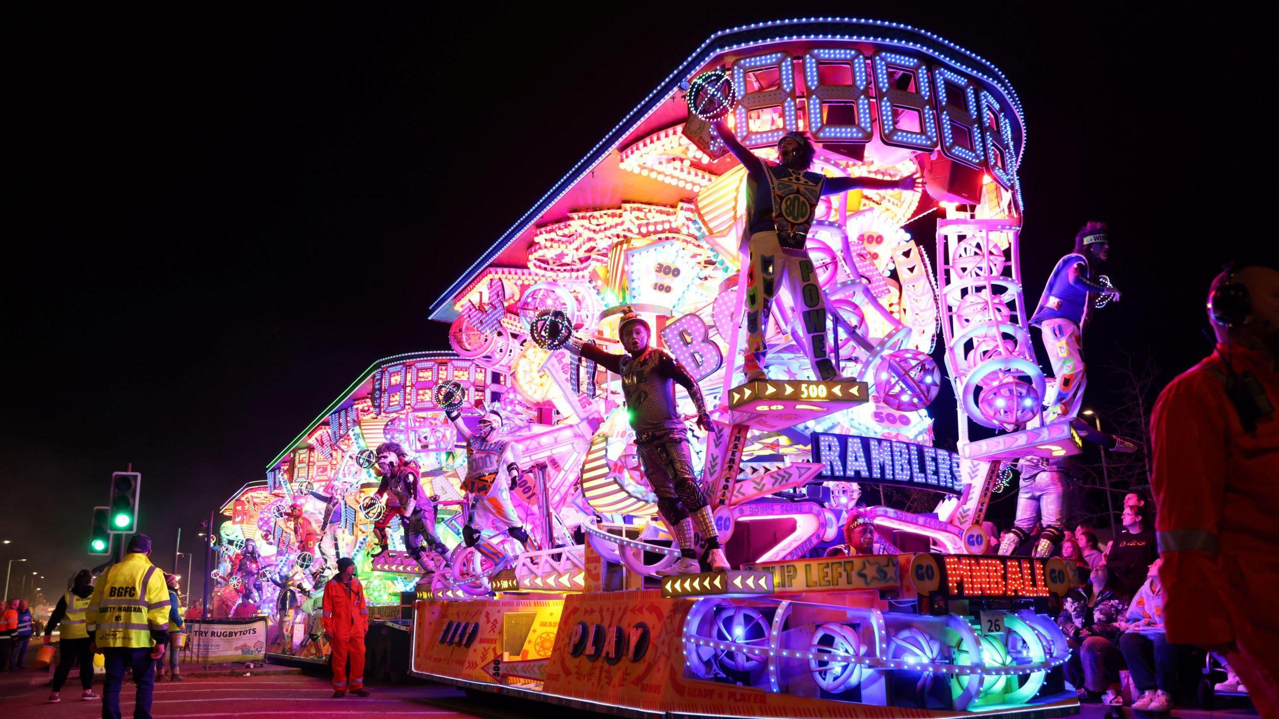 A brightly-lit carnival cart with mostly pink lighting and people hanging off the edge of it